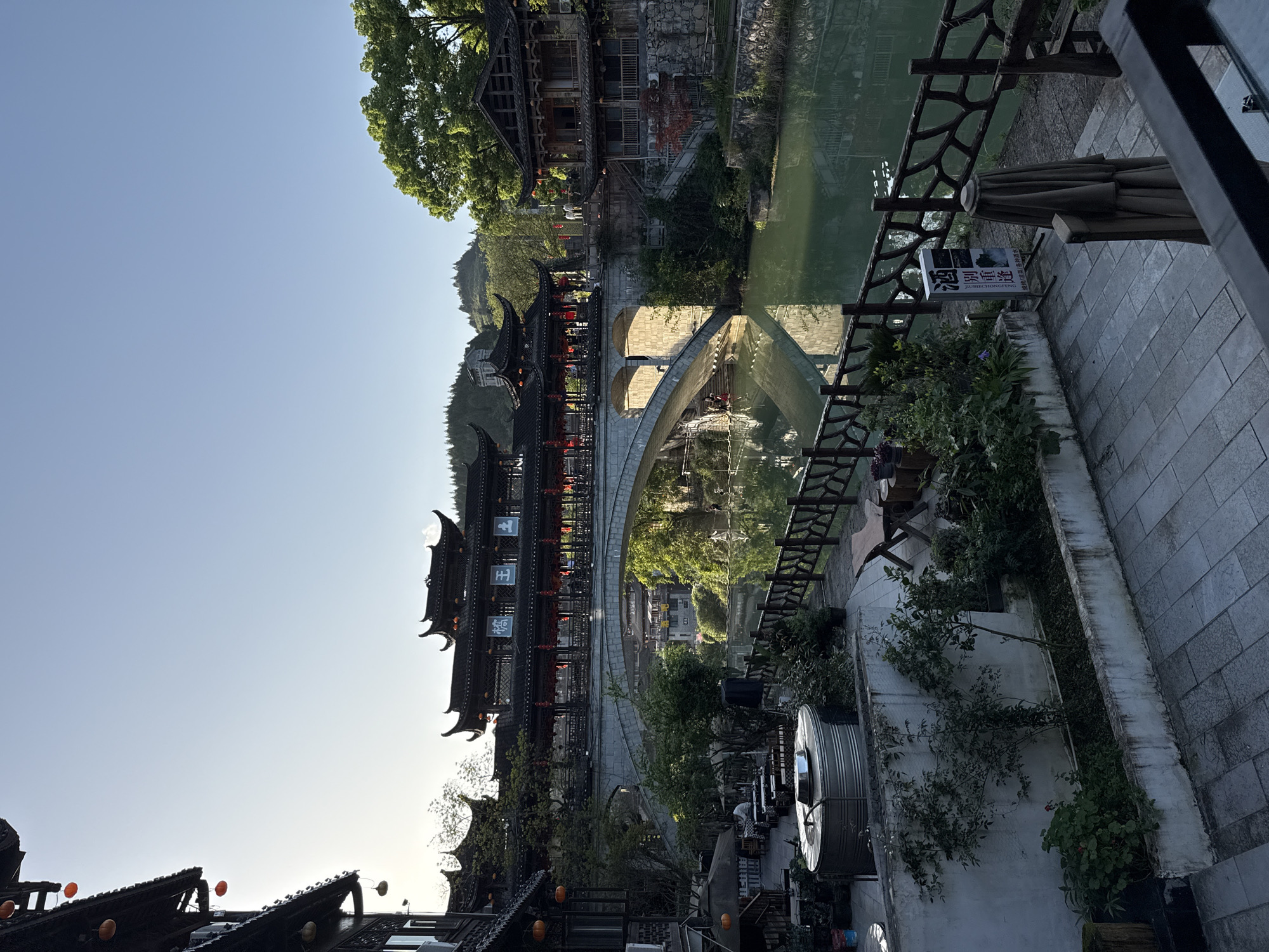 The image presents a view of Furong Ancient Town, China. The subject is a bridge with traditional Chinese architecture built on top, spanning a river. Red lanterns adorn the bridge. The river's surface reflects the bridge and surrounding greenery. On the right side of the image, a stone path or walkway is present with plants and restaurant seating. The foreground shows elements of traditional Chinese architecture with curved roofs. A mountain rises in the background. The sky is clear.