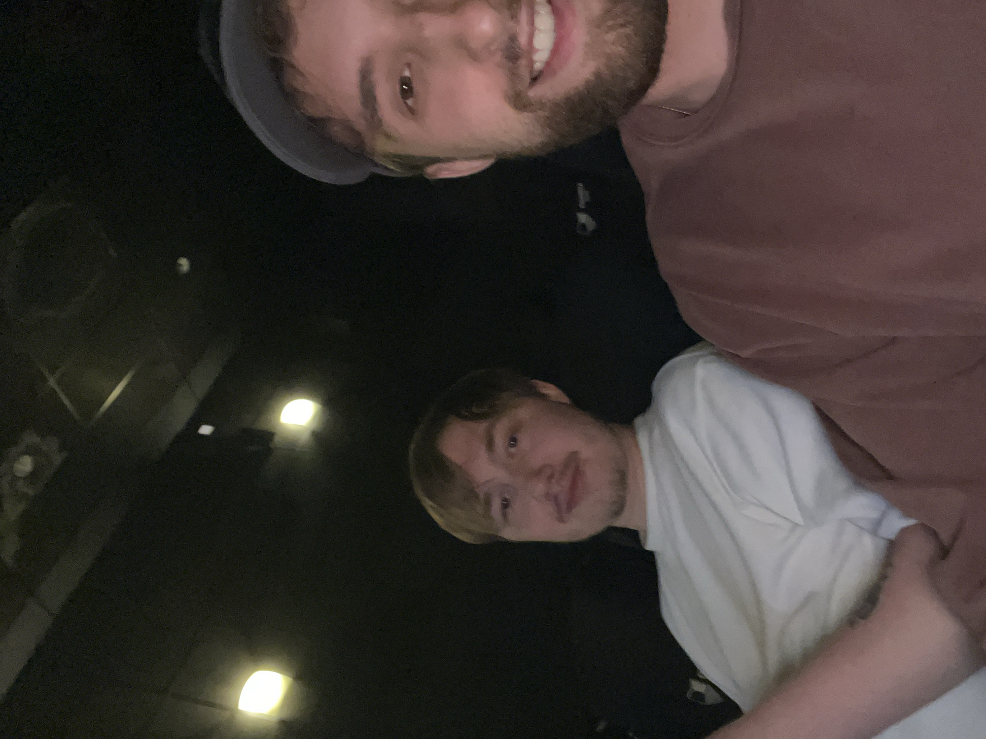 Two young men are visible in the image. One, with a beard and wearing a brown shirt, is smiling. The other is wearing a white shirt. The image seems to be taken at night with a dark background. There are some visible lights and architecture in the background. The image could be anywhere, as nothing distinct identifies it with Krasnoyarsk, Russia.