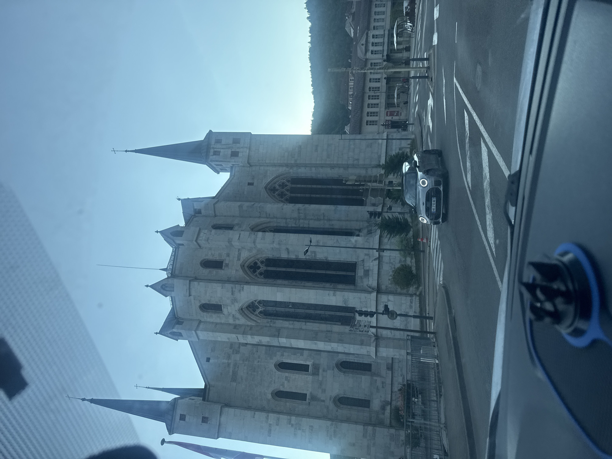 The image depicts a street scene in Saint-Claude, France, with a prominent stone church in the background. The church features tall spires topped with crosses and gothic-style arched windows. In the foreground, a silver Mini Cooper is driving through an intersection. The street is paved, and there are crosswalks visible. Buildings line the street on the right side, and a forested hill rises behind them. The sky is a clear, pale blue, suggesting it is either early morning or late afternoon. The perspective is from inside a vehicle, looking out through the windshield. A car mount for a phone is visible in the lower part of the frame, on the dashboard.