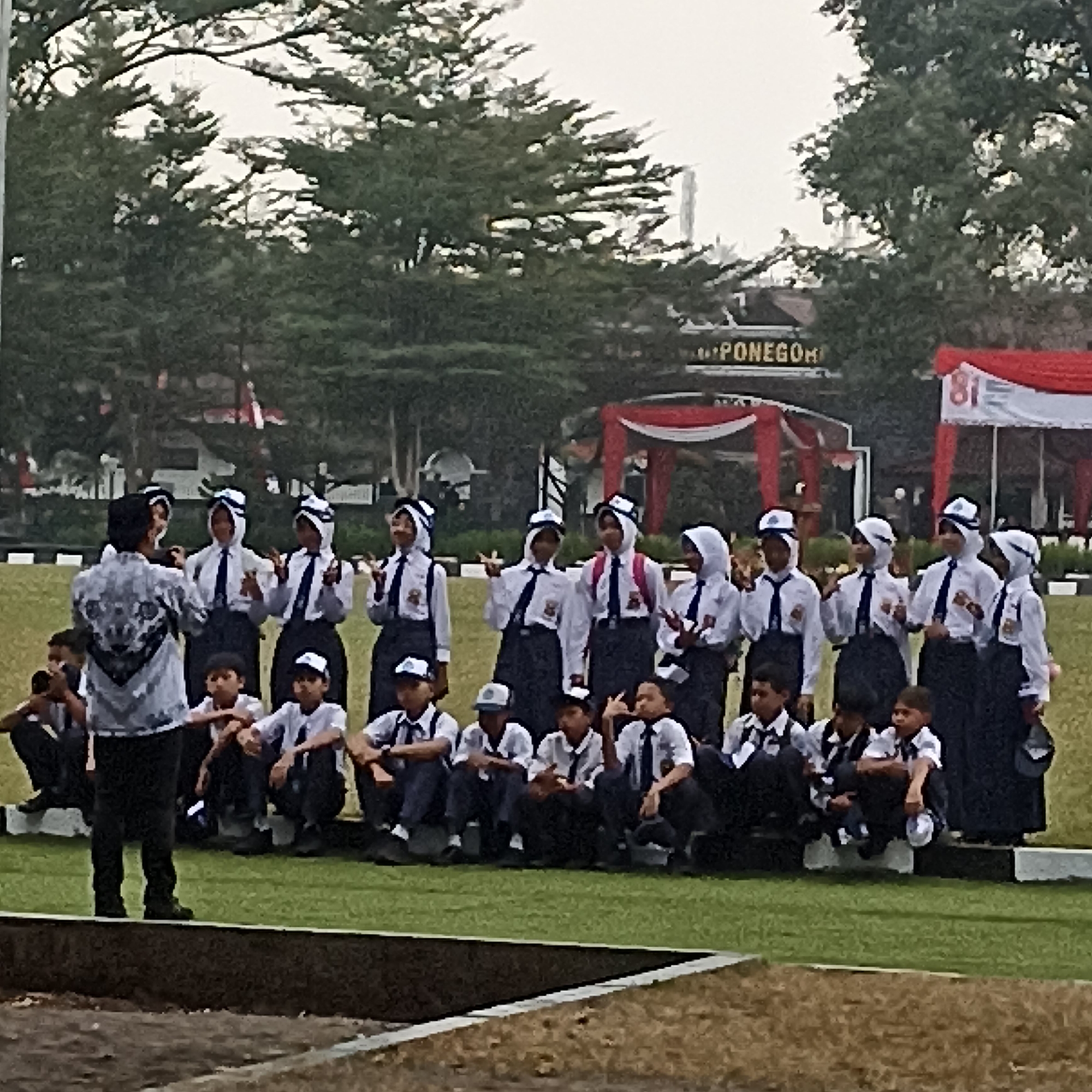 A group of young students in uniform, some wearing hijabs, are posing for a photo outdoors. They are on a grassy field, with trees and a building in the background. Some students are sitting, while others are standing. A man in a patterned shirt is in the foreground, facing away from the camera. The building in the background appears to be decorated with red and white banners, possibly for an event. The overall scene suggests a school event or ceremony. The image quality is somewhat blurry, making it difficult to discern fine details. Based on the context provided in the prompt about Magelang, Indonesia, this scene is likely taking place in that city.