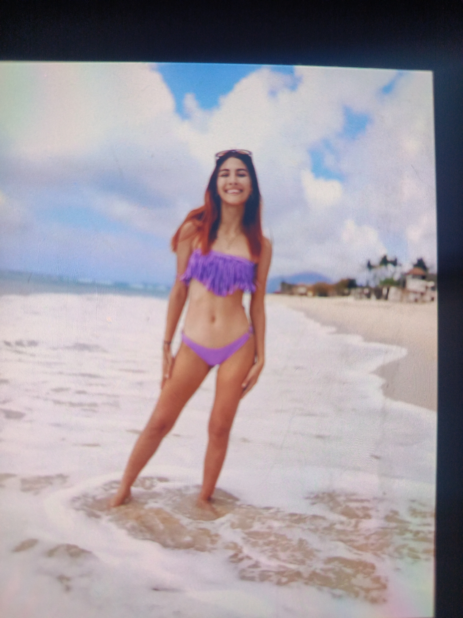 A young woman stands on a beach, partially submerged in gentle waves. She is wearing a purple fringed bikini and sunglasses are perched on her head. The beach appears sandy with white foam from the waves, and the sky has scattered clouds. In the background are blurred buildings and palm trees, consistent with a beach resort area. The scene is likely in or near Juan Griego, Venezuela.