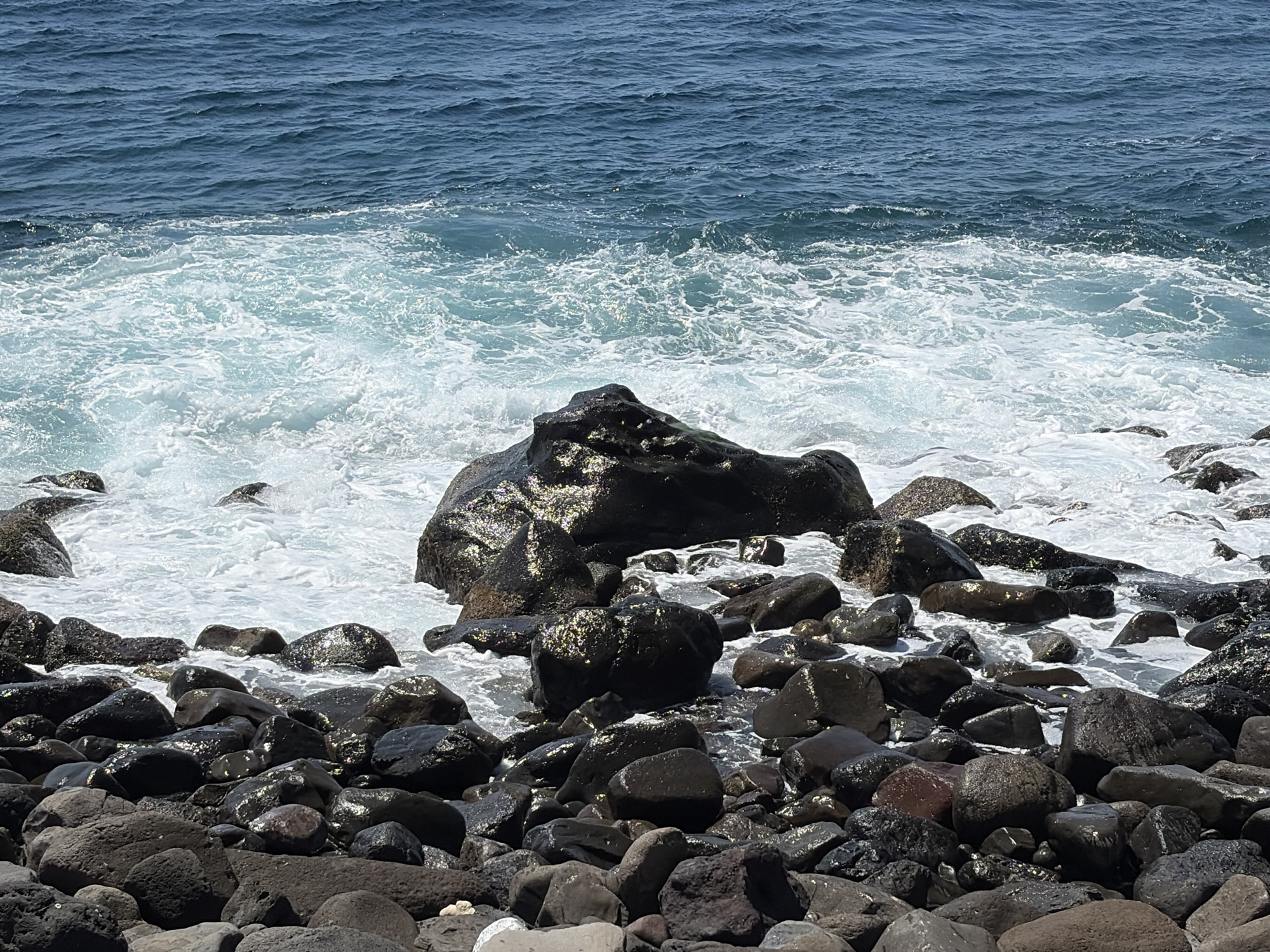 The image depicts a rocky shoreline with the ocean meeting the land. The water is a deep blue, transitioning to lighter shades of turquoise and white as it crashes against the rocks, creating foam and spray. The rocks are dark, wet, and varied in size and shape, some appearing smooth and rounded, others more jagged. The foreground is dominated by a dense collection of these rocks, with some larger boulders visible closer to the water's edge. The lighting suggests it is daytime, with sunlight reflecting off the wet surfaces of the rocks and the water. There are no people or man-made structures visible in the image. The scene is a natural coastal environment.
