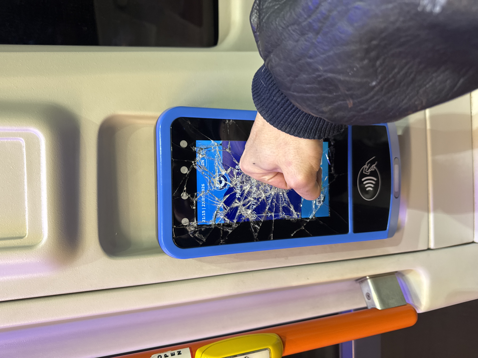 A person wearing a dark blue jacket is punching the screen of a public transportation ticketing machine. The screen is cracked. The ticket machine is mounted to a light tan-colored wall inside a transportation vehicle, such as a bus or train. The date on the screen reads "21:15 | 27.05.2026." The location is possibly in Sofia, Bulgaria, based on the date format (day.month.year). An orange bar and part of a yellow "Open" button are visible at the bottom of the frame.
