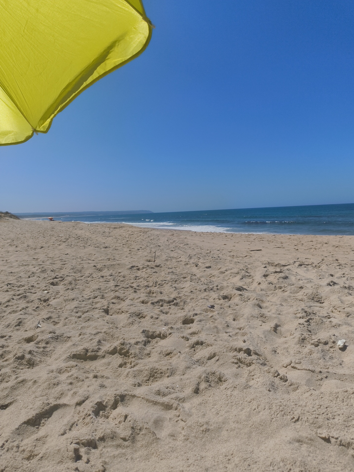 The image depicts a sunny day at a beach in Sesimbra, Portugal. In the foreground, there is a vast expanse of sand with ripples and textures indicating dry conditions. A portion of a bright yellow umbrella is visible in the upper left corner, casting a shadow. In the mid-ground, the sandy beach transitions to the ocean. The sea is calm with gentle waves breaking along the shore. The horizon is clear, showing the blue sky meeting the blue-green water. Further in the distance, along the coastline, low-lying vegetation and a hint of land are visible. The overall scene is one of a serene beach environment under a clear, bright sky.