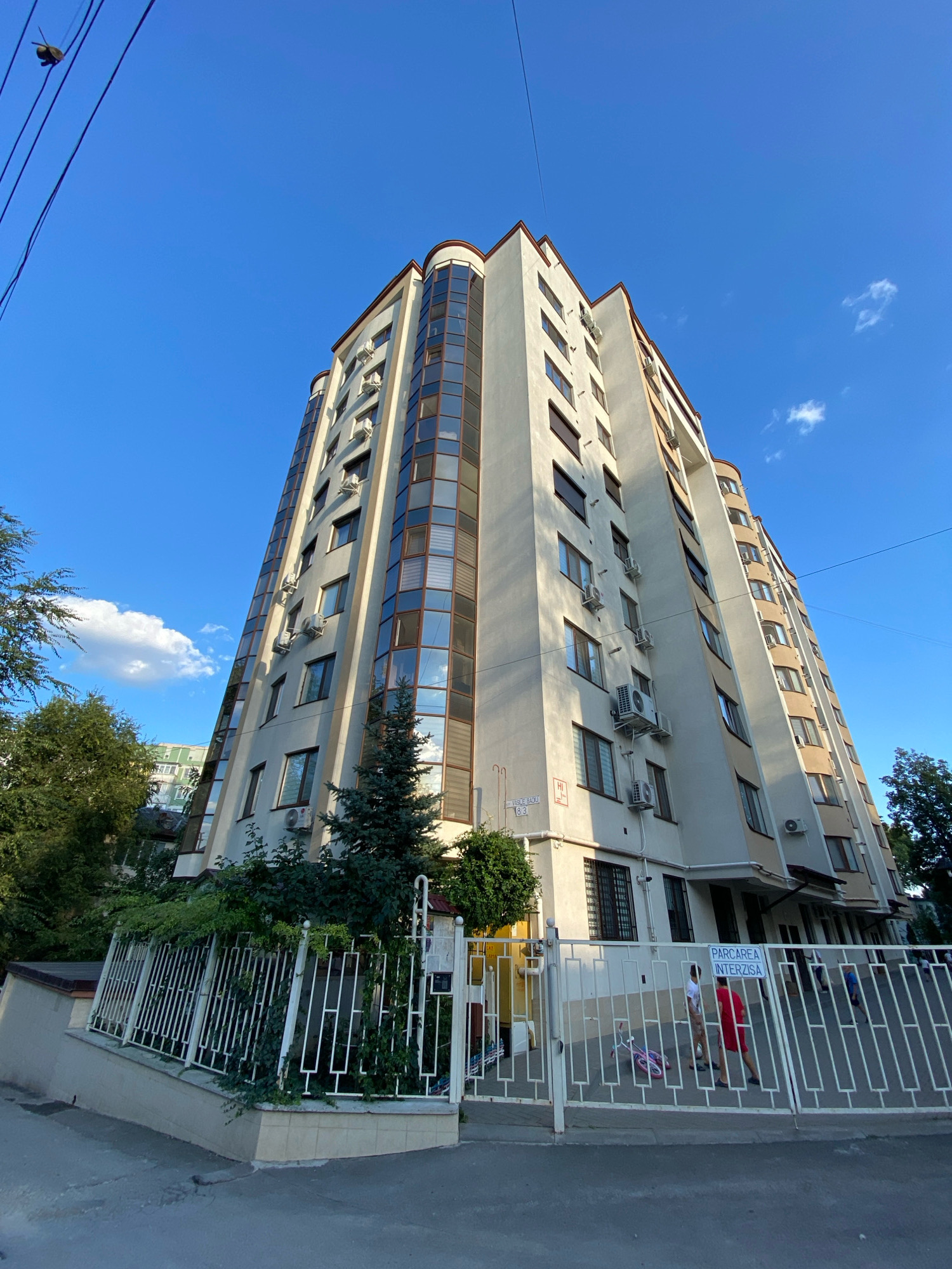 This is a ground-level, wide-angle, outdoor, eye-level shot of a tall, multi-story apartment building with a beige facade and a reddish-brown roof. The building features a curved corner with a wall of windows and numerous air conditioning units mounted on the exterior. A white metal fence encloses the property in the foreground, with a sign that reads "PARCAREA INTERZISA" (Parking Forbidden). Two individuals are visible behind the fence, one wearing a red shirt and the other a white shirt, with a pink bicycle lying on the ground nearby. Trees and other buildings are visible in the background. The sky is blue with scattered white clouds, suggesting it is daytime. The building is located at 83 Vasile Alecsandri Street in Chișinău, Moldova, as indicated by a street sign on the building's facade.