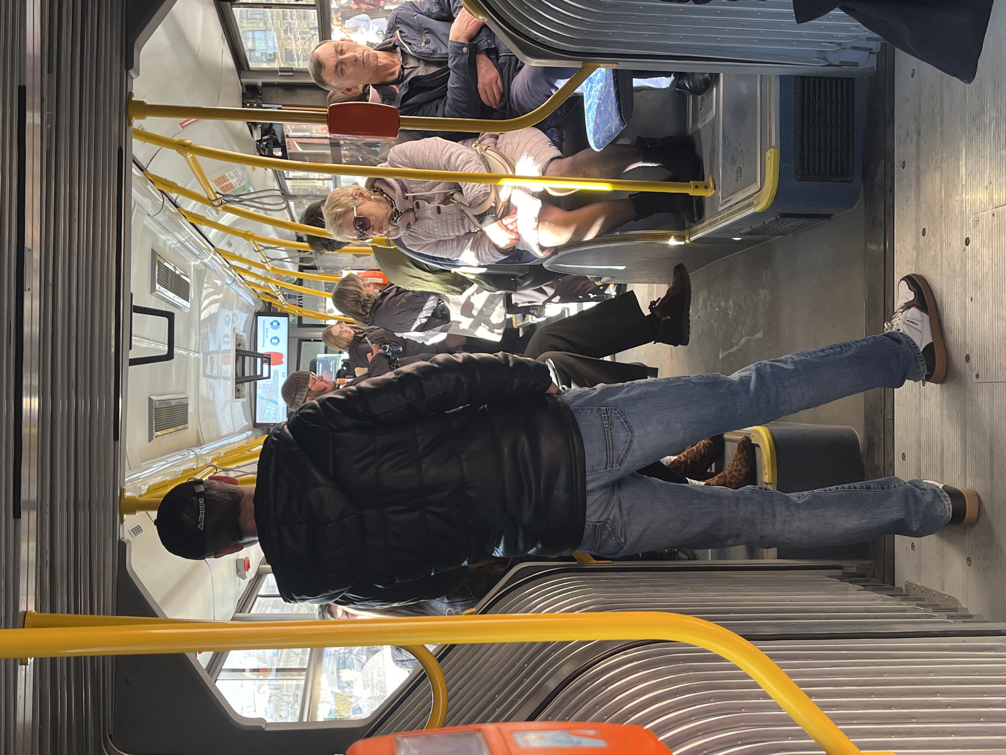 The image displays the interior of a public transit bus in Saint Petersburg, Russia, during daylight. A male individual, viewed from behind, stands prominently in the center of the aisle. He wears a black padded jacket with a hood, blue denim jeans, and a black baseball cap with a white "Kappa" logo. His footwear consists of brown and white sneakers with tan soles.
To the right of the standing male, an older woman is seated, illuminated by sunlight. She wears a light pink quilted coat, dark sunglasses, a patterned scarf, and dark boots, and carries a light-colored shoulder bag. Next to her, an older man is seated, dressed in a dark blue jacket and a grey collared shirt, with his arms crossed. Both are facing generally forward.
Additional passengers are visible throughout the bus interior, some standing and others partially obscured in the background. A woman with light brown hair is seen near a window on the left, possibly interacting with a mobile device.
The bus interior features numerous bright yellow vertical and horizontal grab bars. The walls are light grey with a ribbed texture, and seats are upholstered in a blue patterned fabric. An orange vertical stanchion is present on the far left. A digital display screen is visible near the ceiling on the left, showing digital icons and possibly a route number. The floor is light grey with horizontal grooves. Through the windows, daylight illuminates the scene, revealing classic European-style buildings, other vehicles, and pedestrians outside, consistent with an urban environment like Saint Petersburg.