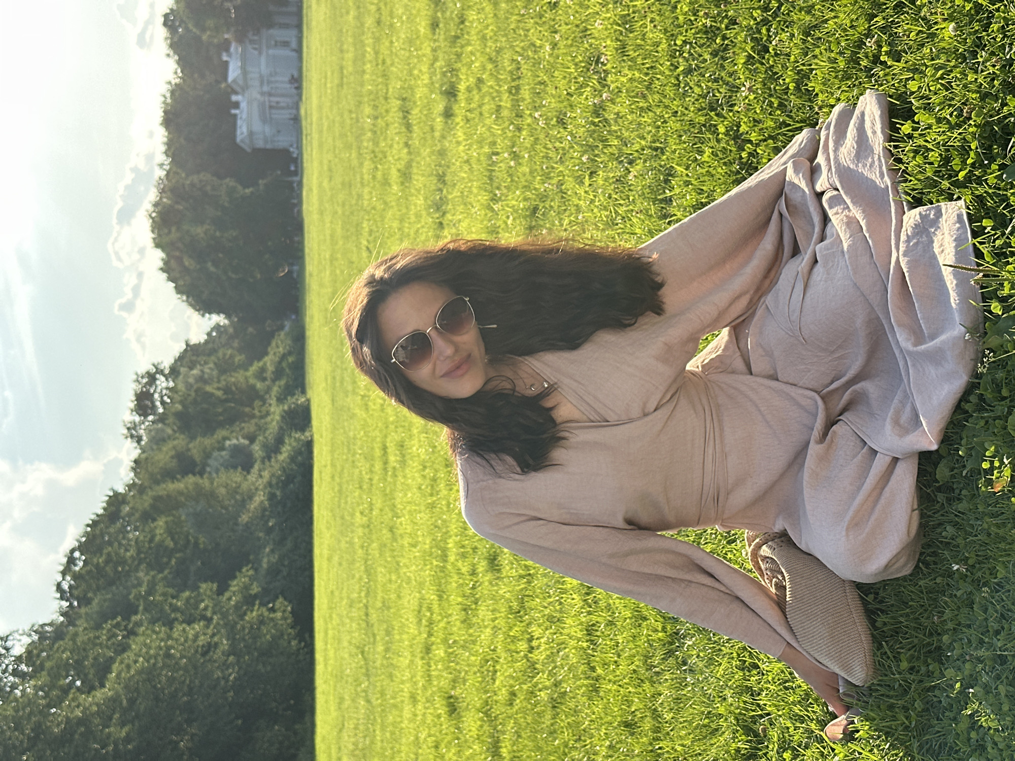 A woman with long, dark, wavy hair and sunglasses is sitting cross-legged on a bright green lawn. She is wearing a light beige, flowy dress with wide sleeves. She is looking towards the camera with a slight smile. To her left, a woven straw bag rests on the grass. In the background, a line of lush green trees borders the lawn, and a pale, classical-style building with columns is visible beyond the trees. The sun is shining, casting a warm glow on the scene, with some clouds in the bright sky. This location is likely the Pavlovsk Park, a part of the State Museum-Reserve Pavlovsk, which is situated in the city of Saint Petersburg, Russia.