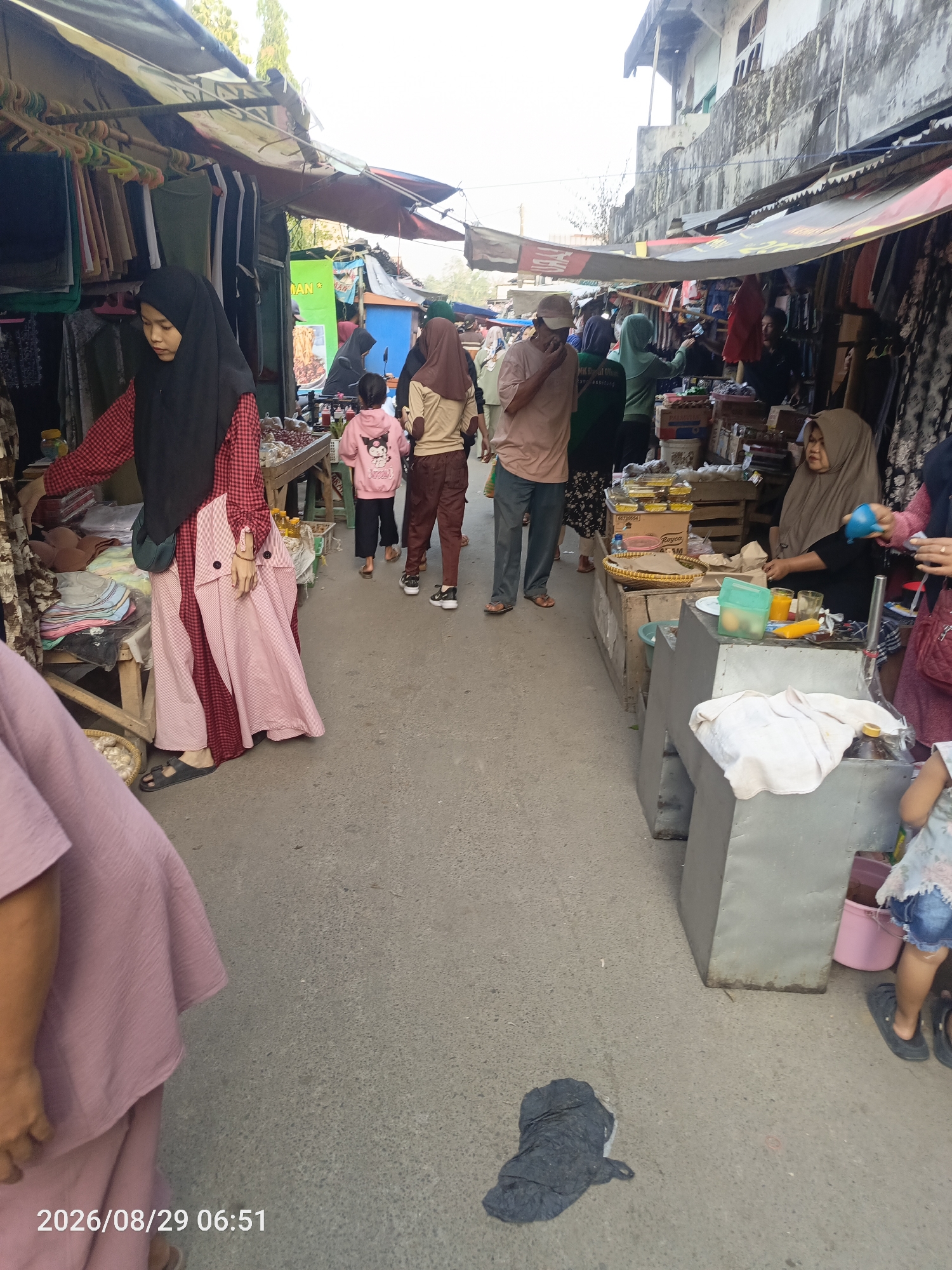 The image depicts a bustling market scene in Sangiang, Indonesia. The market appears to be an outdoor street market with stalls lining both sides of a pathway. The pathway is made of asphalt. Several individuals are visible, engaged in shopping and selling. On the left side of the image, a woman wearing a red and white checkered top and a long pink skirt with white stripes, along with a black hijab, is browsing items at a stall. The stall displays clothing on hangers and stacked fabrics. Further down the pathway, a child in a pink hoodie with a cartoon character is walking. Other individuals in the background are also seen amidst the market stalls. On the right side, a woman wearing a brown hijab is seated behind a stall displaying various items, including food containers and bottles. A child in a floral top and denim shorts is visible on the far right. The stalls are covered with awnings, creating a shaded environment. Various goods, including clothing, food, and household items, are displayed for sale. The overall atmosphere suggests a lively and active marketplace.