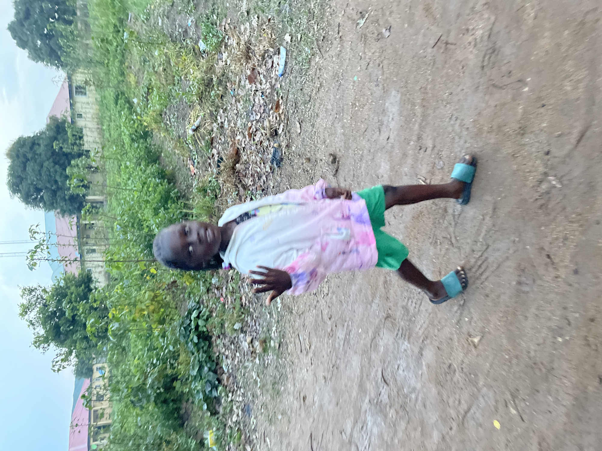 An outdoor, eye-level shot shows a young Black girl from the waist down, walking on a dirt path. She is wearing green shorts, a pink and white hooded jacket, and blue slides. She is looking back over her left shoulder and waving with her right hand. The background is out of focus, but shows green foliage, buildings with red roofs, and debris on the ground. The lighting suggests it is either early morning or late evening. The location is not identifiable as the city of Jos, Nigeria.