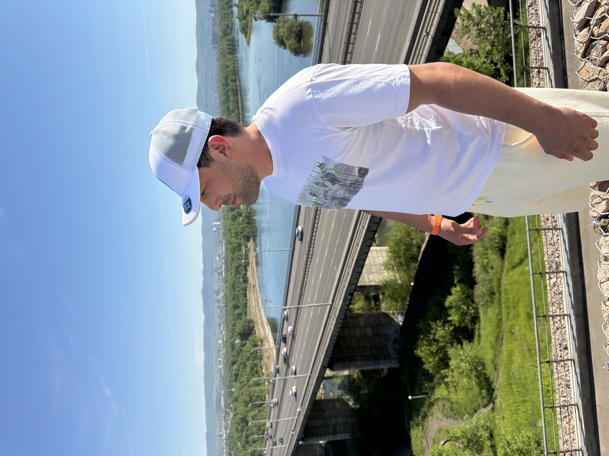 A man is depicted in a right profile, positioned at an elevated viewpoint overlooking the Yenisei River and the city of Krasnoyarsk, Russia. He wears a white trucker cap with a light blue mesh back, a white short-sleeved t-shirt featuring a dark graphic design of palm trees and text, and light cream-colored shorts. An orange wristband is visible on his left wrist. His facial hair includes a short beard and stubble.
In the mid-ground, a multi-lane concrete highway bridge, identifiable as the Oktyabrsky Bridge, spans the wide, blue-green Yenisei River. Several vehicles, including cars and vans, are visible in motion on the bridge. The river shows a distinct bend with lush green trees and foliage along its banks and on an island formation.
The background reveals the cityscape of Krasnoyarsk with numerous buildings, extending towards distant green hills or mountains under a clear, bright blue sky with a few faint wisps of clouds.
In the immediate foreground, the man stands behind a metal railing, which rests on a concrete structure with visible gabion baskets (wire cages filled with rocks). Below this barrier, a steep grassy slope with dirt paths and abundant green vegetation descends. The lighting suggests a bright, sunny day.