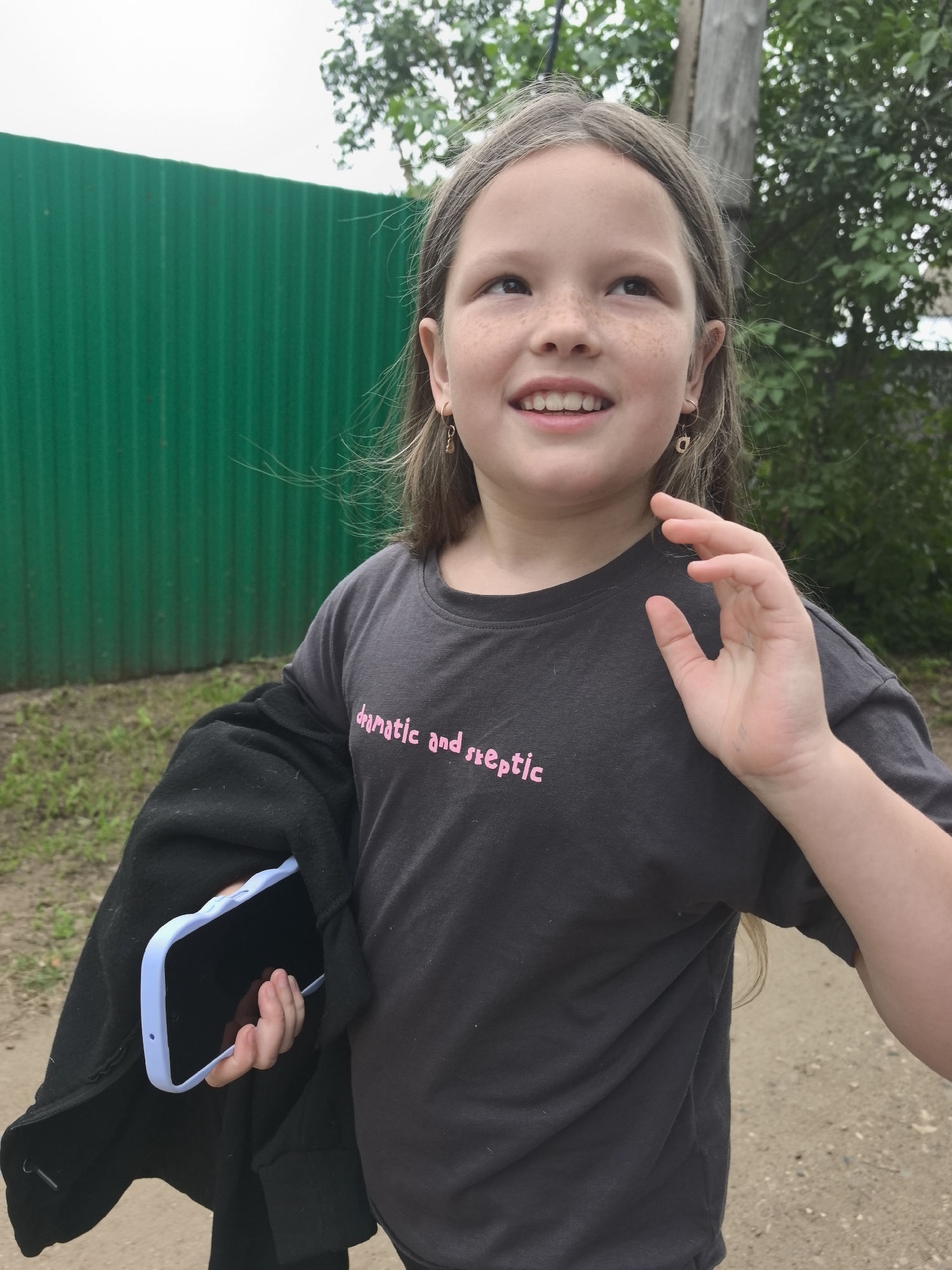 This is a medium shot, eye-level, outdoor photograph of a young girl with blonde hair and freckles. She is looking upwards and to her right with a happy expression, smiling with her mouth open. She is wearing a dark grey t-shirt with pink text that reads "dramatic and skeptic". She has small gold hoop earrings with a charm. She is holding a black jacket over her left arm, with a light blue phone case peeking out from underneath. Her right hand is raised, with her fingers slightly curled.
The setting appears to be outdoors in a rural or suburban area, possibly Noviy, Russia. Behind her is a bright green corrugated metal fence. To the right of the girl, there is a wooden pole and lush green trees and foliage. The ground is a dirt path with small pebbles. The lighting suggests it is daytime, possibly overcast given the bright, diffused light and the color of the sky. The overall mood is cheerful and casual.