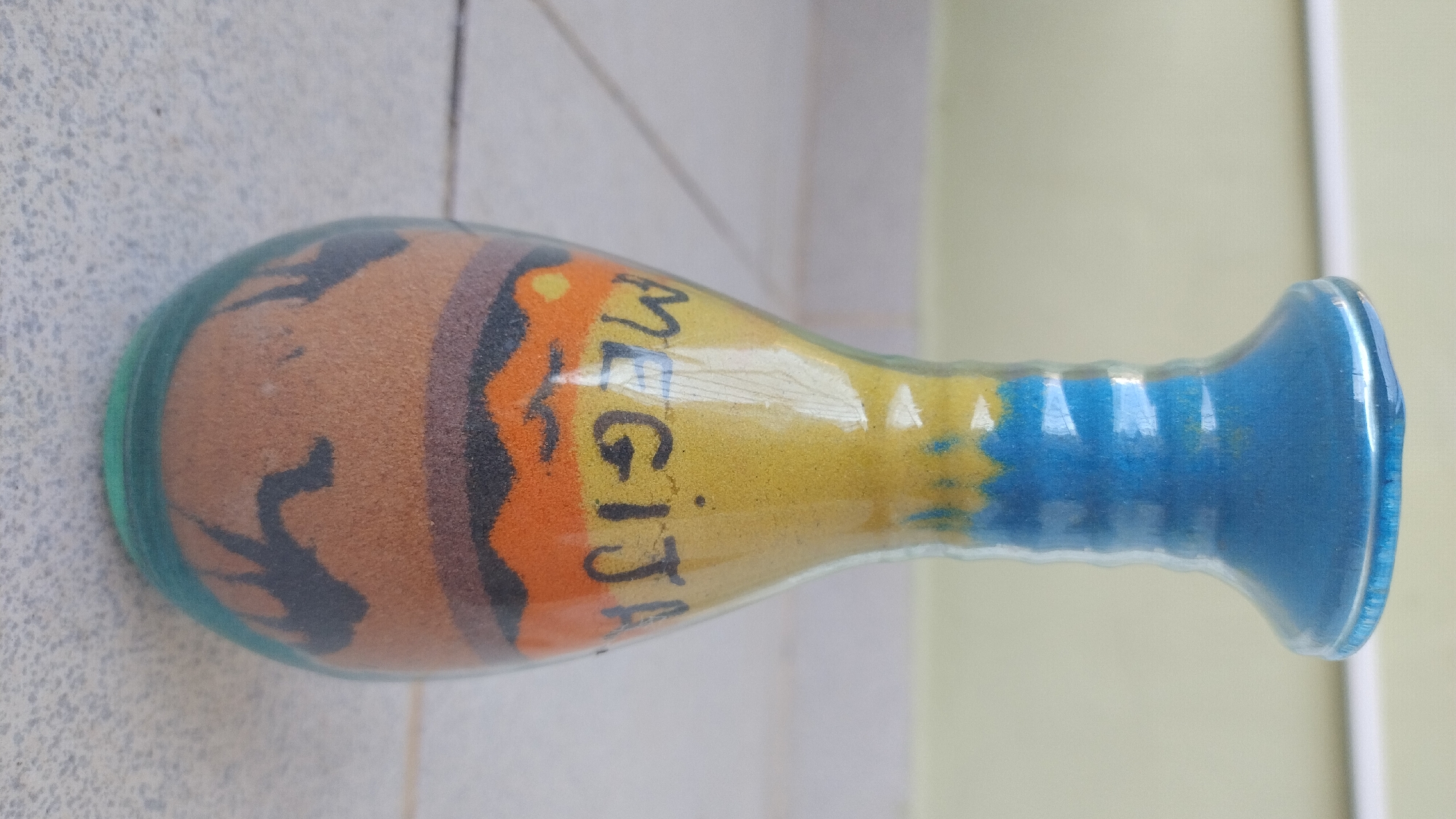 A vase made of layered colored sand displays a desert scene with camels and a sunset. The vase is primarily yellow and blue, with a band of orange and black sand depicting rolling hills and a sun. The name "MEGIJA" is written in black in the upper portion of the sand art. The vase has a narrow neck and a wider body, and it rests on a surface that appears to be tiled. The background is a plain, light-colored wall. The image does not provide any information about the city of Perea, Greece.