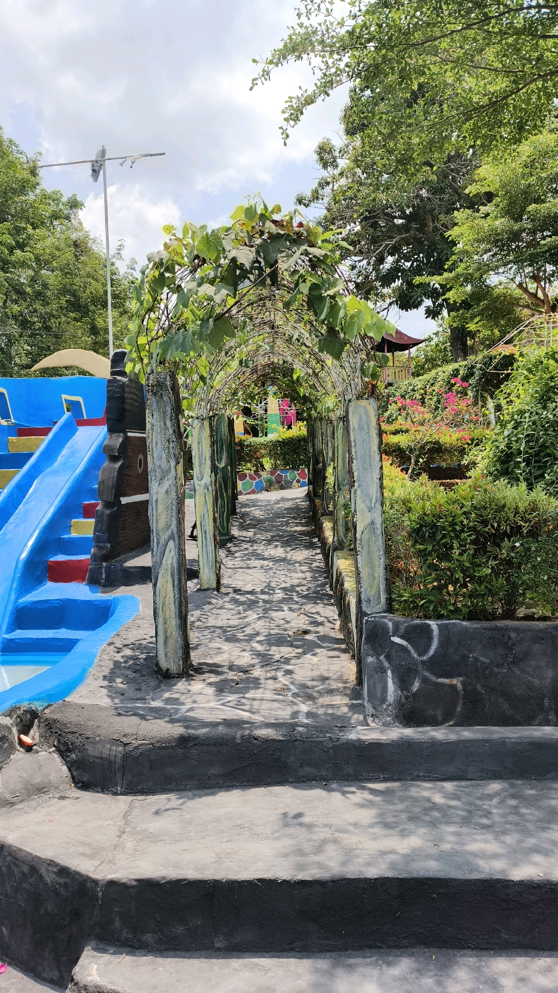 The image captures a pathway covered by an arched trellis adorned with green vines, leading into a garden area. The pathway is paved with stone tiles, and shadows from the trellis and overhead trees create a dappled effect on the ground. On the left side of the pathway, a vibrant blue slide is visible, part of what appears to be a recreational facility. The garden beyond the trellis features lush greenery, flowering plants with pink blossoms, and a small, red-roofed pavilion. The scene is outdoors, under a bright sky with scattered clouds, and appears to be located in a tropical or subtropical environment. The overall impression is one of a serene and picturesque garden setting, possibly a park or resort.