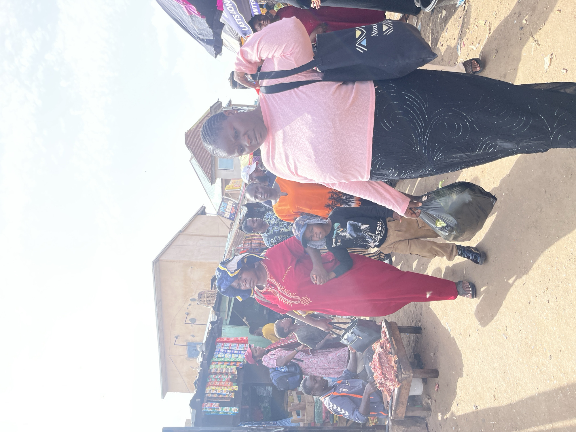 This is a photograph taken in Rafin Par, Nigeria, during the daytime under bright, possibly overcast skies. The scene depicts a bustling outdoor market with several people engaged in various activities.
In the foreground, a woman in a red dress and blue headscarf is the central focus. She is looking towards the camera with a neutral expression. To her right, a child with a grey hood is looking away. Behind them, a woman in a light pink top and a patterned black skirt is holding a black bag. Another woman in an orange shirt is visible behind her. Further in the background, a person in an orange and blue jacket is standing near a table laden with raw meat. There are stalls with shelves stocked with colorful products, suggesting food and other goods are being sold. Several other individuals are present in the crowd, some carrying bags.
The ground is dusty and appears to be a dirt path. The overall atmosphere suggests a lively market day. Visible text includes "LASTS LON" on a black banner in the upper left portion of the image and "ALUMNI" on the black bag.