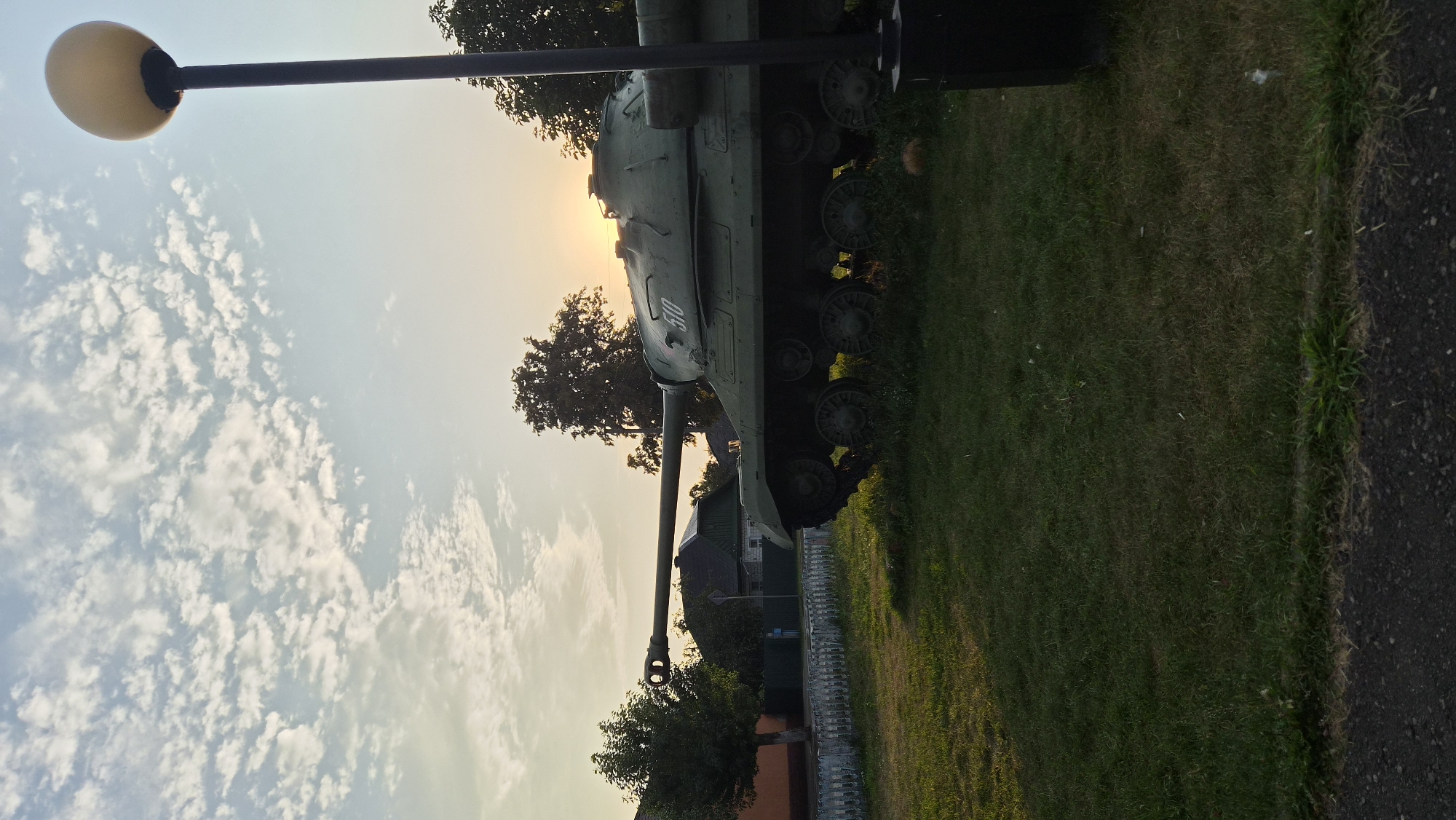 A military tank is parked on a grassy area in front of a building and trees, with a lamp post to the right. The sun is setting behind the tank, casting a warm glow over the scene. The tank is green and appears to be an outdoor monument. The ground is covered in green grass, and there is a white picket fence in the background. The sky is blue with scattered white clouds. The image is taken from a low angle, looking up at the tank. The overall impression is one of peace and tranquility, despite the presence of the military vehicle. The location is Парыцкі сельскі Савет, Belarus.