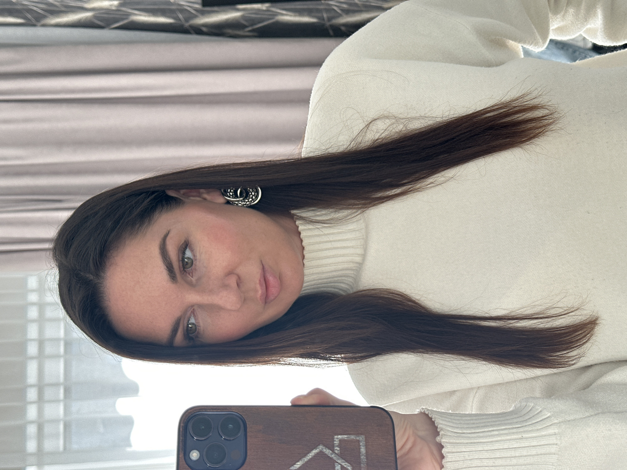 A woman with long, dark brown hair and light green eyes is taking a selfie with her iPhone. She is wearing a white turtleneck sweater and silver earrings. The background is blurry, but it appears to be a room with beige curtains. The location cannot be determined as Chaikovsky, Russia, from the given image.