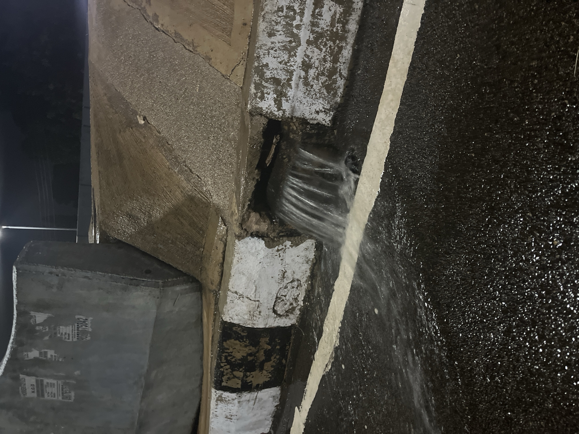 The image captures a street-level view of a broken concrete drain opening on a road in Jos, Nigeria at night. Water is gushing out of the drain, flowing across the wet asphalt road. The road is marked with a white line, which is partially submerged by the water. The concrete barrier above the drain is damaged, with some white and black painted markings visible on it. The overall scene suggests a drainage issue, possibly due to heavy rainfall or a damaged infrastructure, contributing to a wet and somewhat neglected urban environment.
