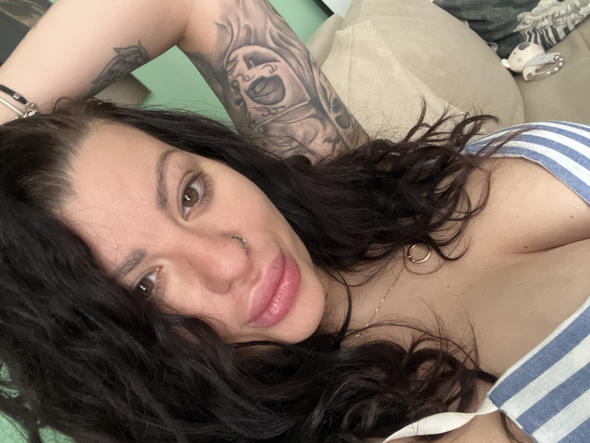 A selfie-style close-up depicts a woman with dark, wavy hair. She is lying down, with her left arm raised above her head, revealing tattoos. The woman has light skin, brown eyes, and a nose ring. She's wearing a striped blue and white garment. A gold necklace is visible on her chest. The background includes a light-colored pillow or cushion and a baby toy with a rattle. The location is not identifiable as Plovdiv, Bulgaria from the image provided.