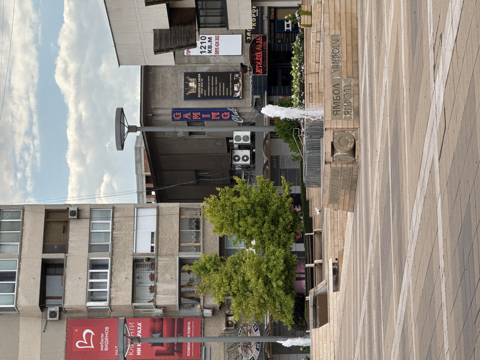 The image depicts an outdoor public space in Yambol, Bulgaria, featuring a modern plaza with paved walkways and a central fountain. In the background, a multi-story building with a concrete facade dominates the scene, displaying various advertisements, including a large red banner for "MEBELI VIDENOV" and another for "GAMING Club". Adjacent to this structure is another building with a white facade. The plaza itself has tiered stone seating areas and planters with greenery. A white dove is visible in mid-flight near a bench. The sky above is partly cloudy. The text "Ямбол" (Yambol) is inscribed on a stone monument in the foreground, confirming the location.