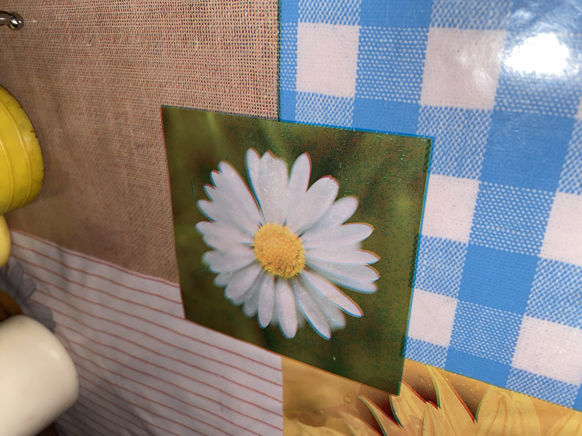 The image shows a close-up shot of a surface displaying several different patterns. A tan fabric with a small, woven texture is partially visible on the left side. Adjacent to it, a white fabric with narrow red stripes runs horizontally. In the center of the image, there is a square-shaped image of a white daisy with a yellow center set against a green background. To the right of the daisy image, a blue and white checkered pattern fills a portion of the upper-right side of the image. Beneath the daisy and checkered patterns, a yellow color with sunflower-like shapes is visible at the bottom of the image. Parts of what seems to be yellow items are present on the left. The exact location in Tinaquillo, Venezuela is unable to be determined from the image.