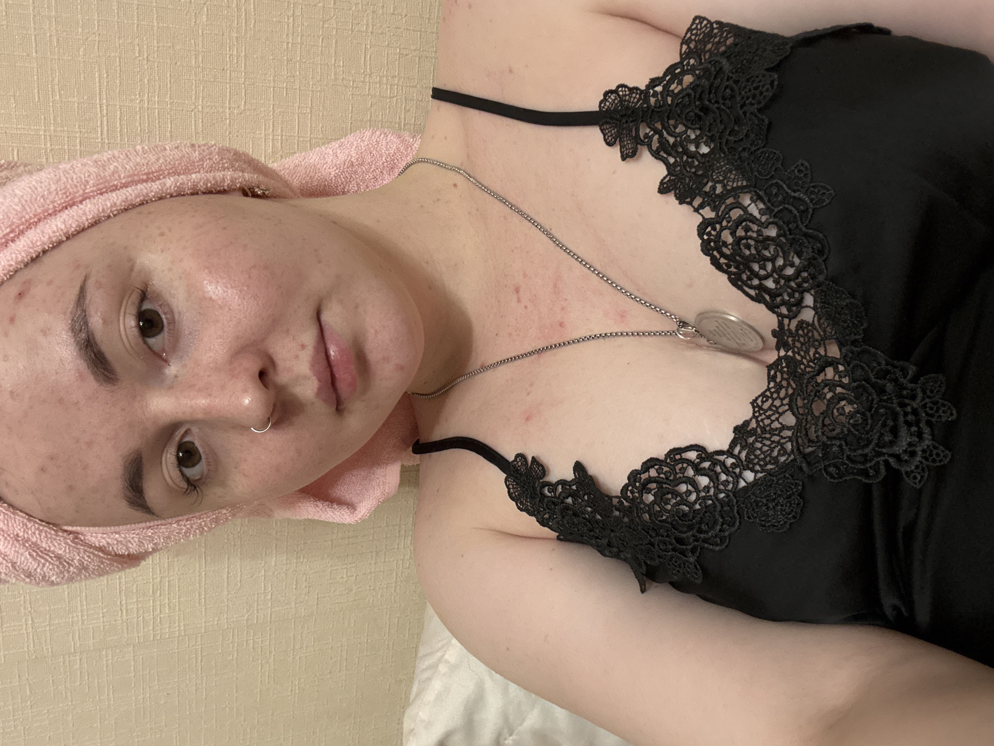 A young woman is lying down, facing the camera at an angle. She has a pink towel wrapped around her head, and is wearing a black satin camisole with black lace trim. She has a silver necklace with a circular pendant. The background appears to be a wall and bedding. There is no indication of a location in Izhevsk, Russia.