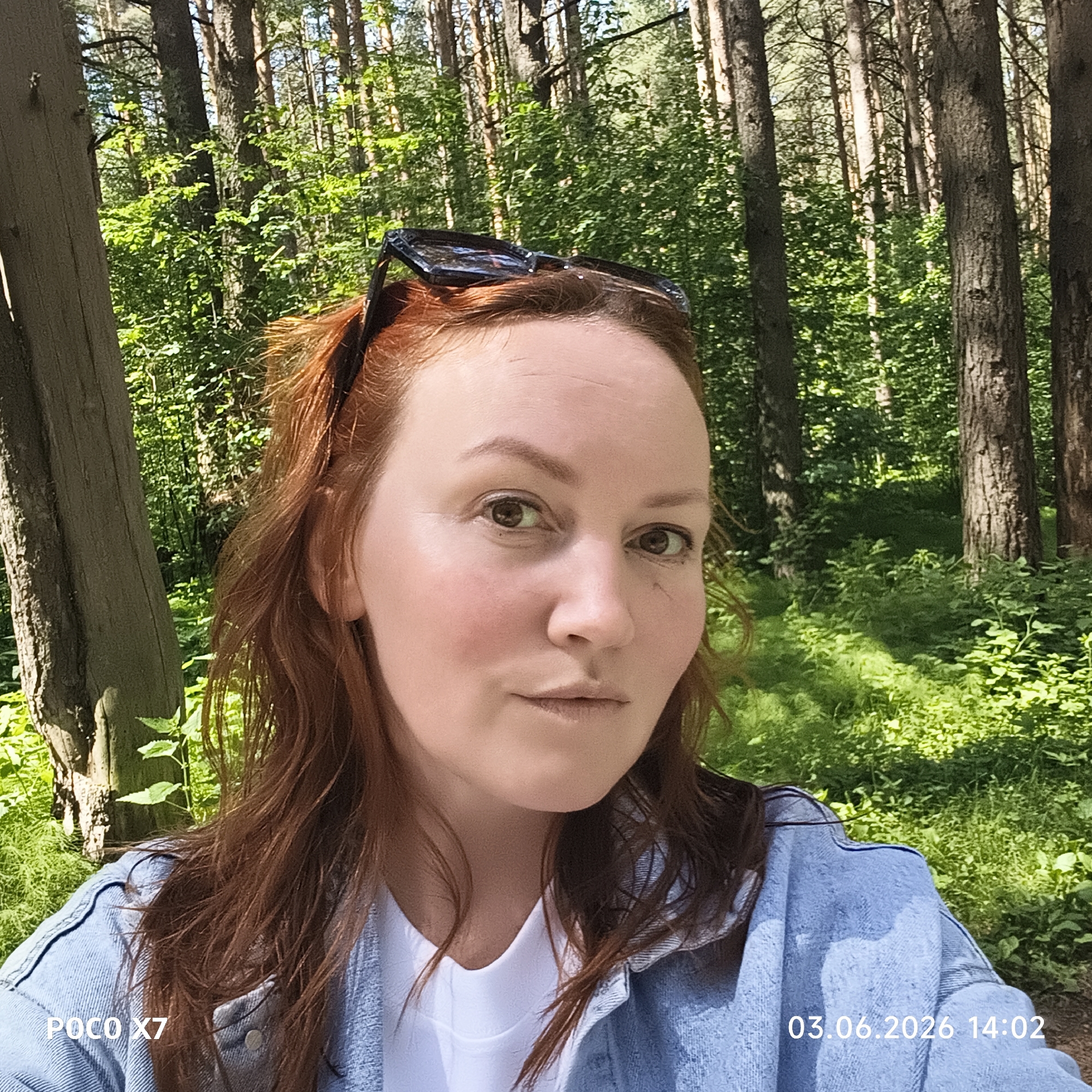 The image captures a close-up selfie of a woman in a lush forest setting, likely in Chaikovsky, Russia. She has reddish-brown hair, dark eyes, and a neutral, contemplative expression as she looks directly at the camera. Dark sunglasses are perched on top of her head, and she is dressed in a light blue denim jacket over a white t-shirt.
The background is dominated by a dense forest, characterized by numerous tall coniferous trees, possibly pines, with visible bark textures. The forest floor and surrounding areas are rich with vibrant green foliage and undergrowth, indicating a healthy and verdant natural environment. The scene is brightly illuminated by natural daylight, suggesting a clear and sunny afternoon.
The overall atmosphere conveys a sense of calm and being immersed in nature. Watermarks in the image indicate the capture details: "POCO X7" is visible in the bottom left, denoting the camera device, and "03.06.2026 14:02" appears in the bottom right, showing the date and time the photograph was taken.