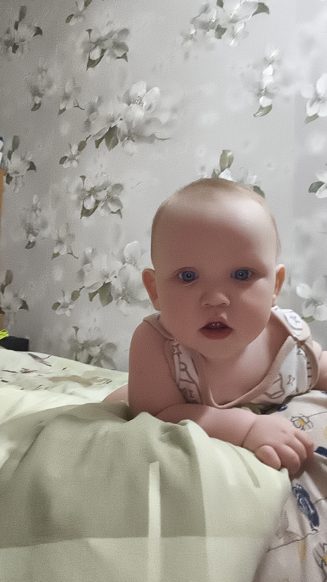 A baby lies on its stomach on a bed. The baby has fair skin, blue eyes, and is wearing a light-colored sleeveless outfit. The bed has a light green blanket or sheet on it. The background is a wall with a floral pattern of white flowers and green leaves. The city of Chaikovsky, Russia, is not discernible from the image.