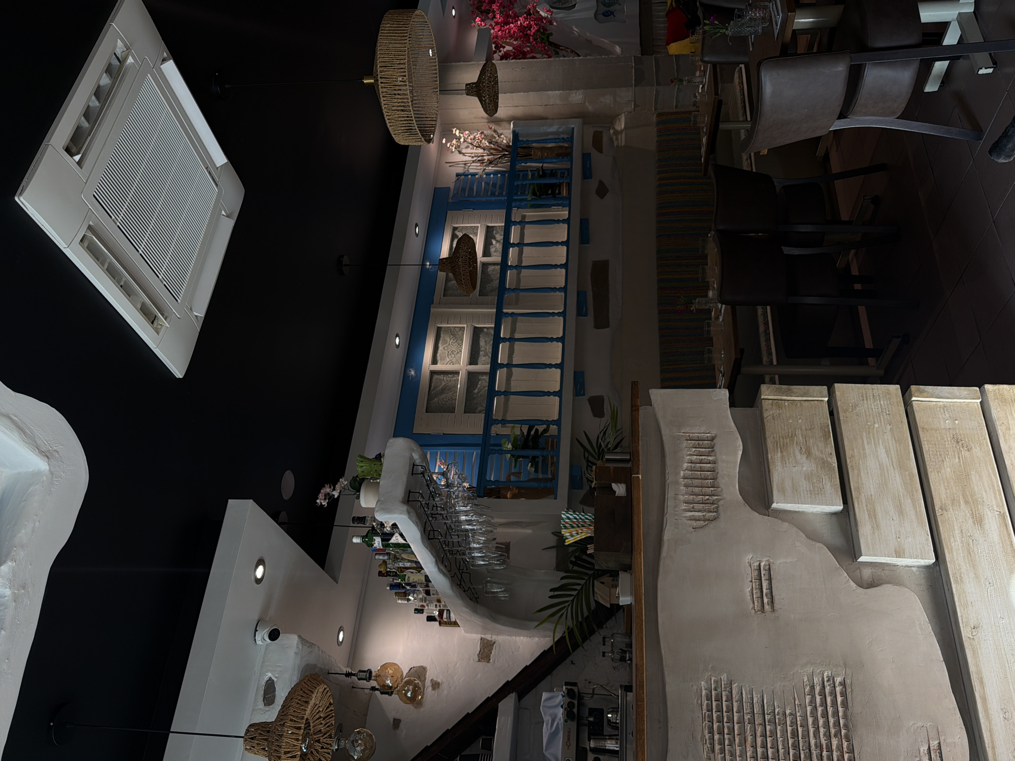 The image depicts the interior of a restaurant. The overall aesthetic appears to be inspired by Mediterranean or Greek island architecture, with whitewashed textured walls, a blue and white balcony with decorative railings, and woven pendant lights.
In the foreground, a curved bar area is visible with textured plaster and wooden elements. On the bar, there is a coffee machine, glassware hanging from a rack, and a collection of bottles on a raised shelf behind the bar. There are also decorative plants and straws in a wooden holder on the bar.
The seating area in the background features tables and chairs. The chairs are dark brown, possibly upholstered. The tables are set with glasses and what appear to be small vases with flowers. A long, cushioned bench with colorful stripes runs along the back wall.
The ceiling is black, contrasting with the white architectural elements. A large, modern air conditioning unit is installed in the ceiling. Several pendant lights, made of woven natural materials, hang from the ceiling. There is also a security camera mounted on one of the white architectural beams.
The lighting in the restaurant is somewhat dim, with spotlights and the pendant lights creating pools of illumination. The color palette is dominated by white, blue, and natural wood tones, with pops of color from the seating and decorative elements.
Without additional information or context, it is impossible to determine the specific location of this restaurant within the city of London, United Kingdom, or to identify any people or specific actions. The image captures a still moment within the restaurant's interior.