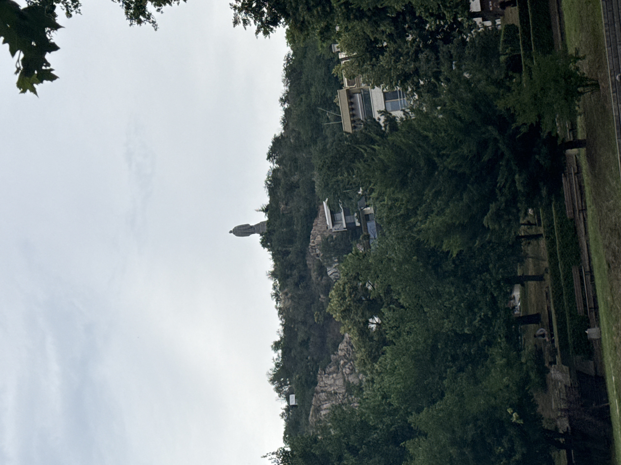 The image shows a statue of a person standing on a hill, surrounded by trees and buildings. The statue is in the distance and appears to be made of stone or bronze. The hill is covered in rocks and vegetation, and there are several buildings on the hillside, including a white building with windows. The statue is a prominent feature of the landscape. The image is taken from a low angle, looking up at the statue and the hill. The sky is overcast, and the weather appears to be mild. The scene is peaceful and serene. The statue is a symbol of strength and resilience. It is a reminder of the history and culture of Plovdiv, Bulgaria.