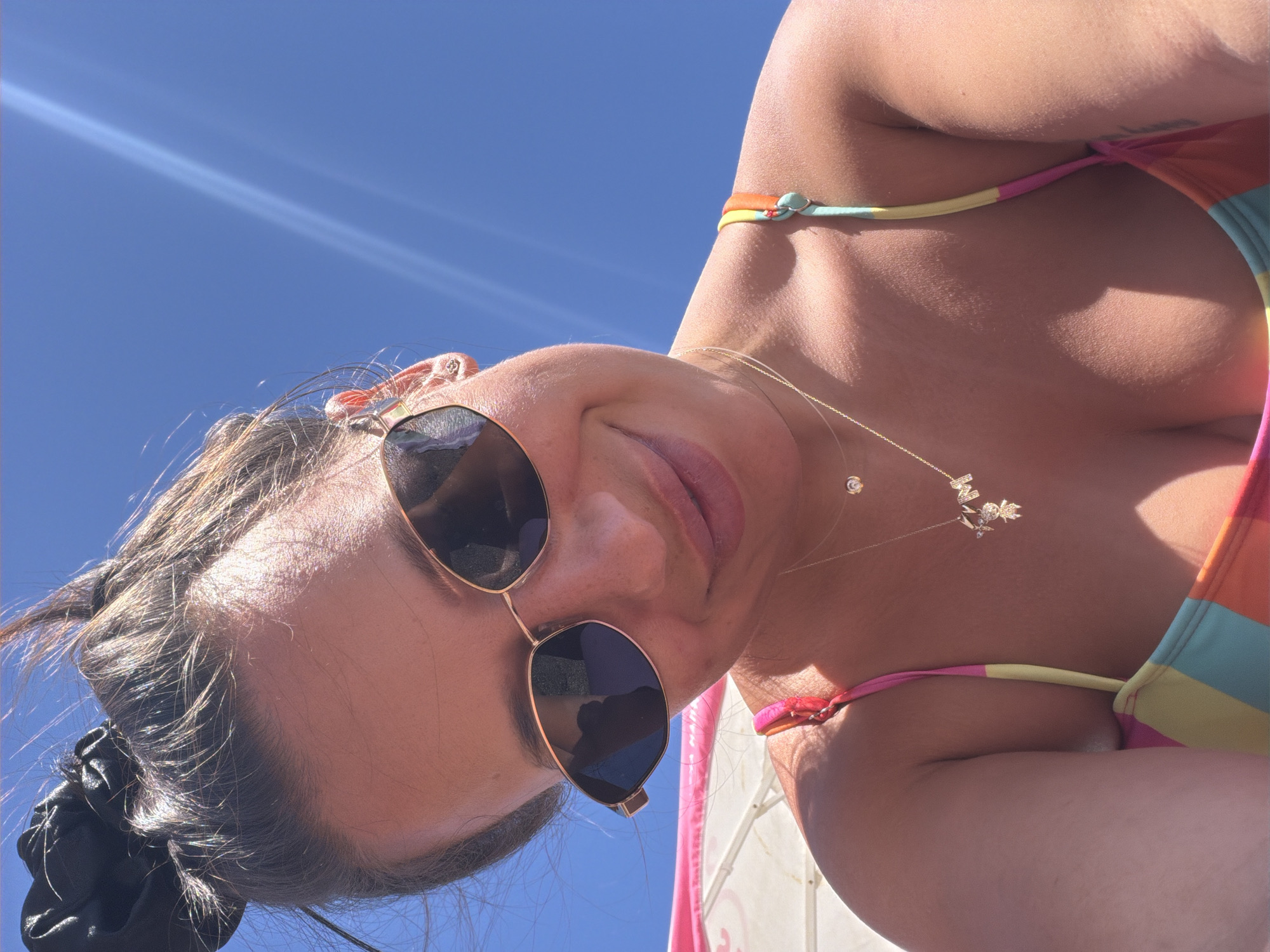 A woman is taking a selfie outdoors on a sunny day, against a bright blue sky. She is wearing sunglasses and a colorful bikini. A beach umbrella is visible in the background, suggesting she is at the beach. The sun is shining brightly, creating lens flares in the image. The overall scene is casual and relaxed, likely a vacation setting. The location is not specified to be in the city of Burgas, Bulgaria.