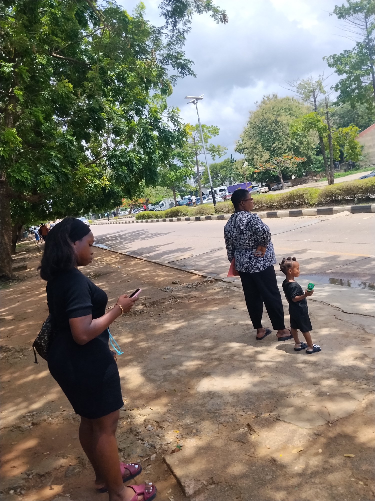 The image shows three people on a sidewalk in Lagos, Nigeria. On the left, a woman in a short black dress is looking at her phone. She is wearing sandals and has a small backpack. Further down the sidewalk, a woman wearing a patterned blouse and black pants stands next to a young child, both looking towards the street. The child is holding a small green object. The sidewalk is made of dirt and has patches of shade from the trees lining the street. A road with cars and a median with bushes can be seen in the background. A street light is visible in the distance. The sky is partly cloudy.