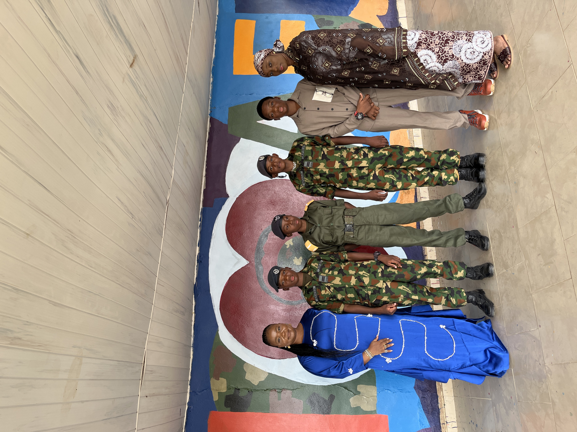 In Bar Jirgi Summa, Nigeria, a group of six stands in front of a colorful mural featuring a large heart and abstract shapes. On the left, a woman in a long, blue satin dress with white bead accents poses with her hand on her hip. Next to her are three young men in military uniforms, two in camouflage and one in olive green. The camouflage uniforms feature patches with names and insignia. The third man from the left wears a cap and has "APOLLO" written on his uniform. The fourth person, a young man in a light brown suit, stands with his hands clasped. On the far right, a woman in a brown patterned dress and headscarf stands with her arms crossed. The mural is painted on a wall, and above it, a white, slatted ceiling with some stains is visible. The ground beneath them is tiled.