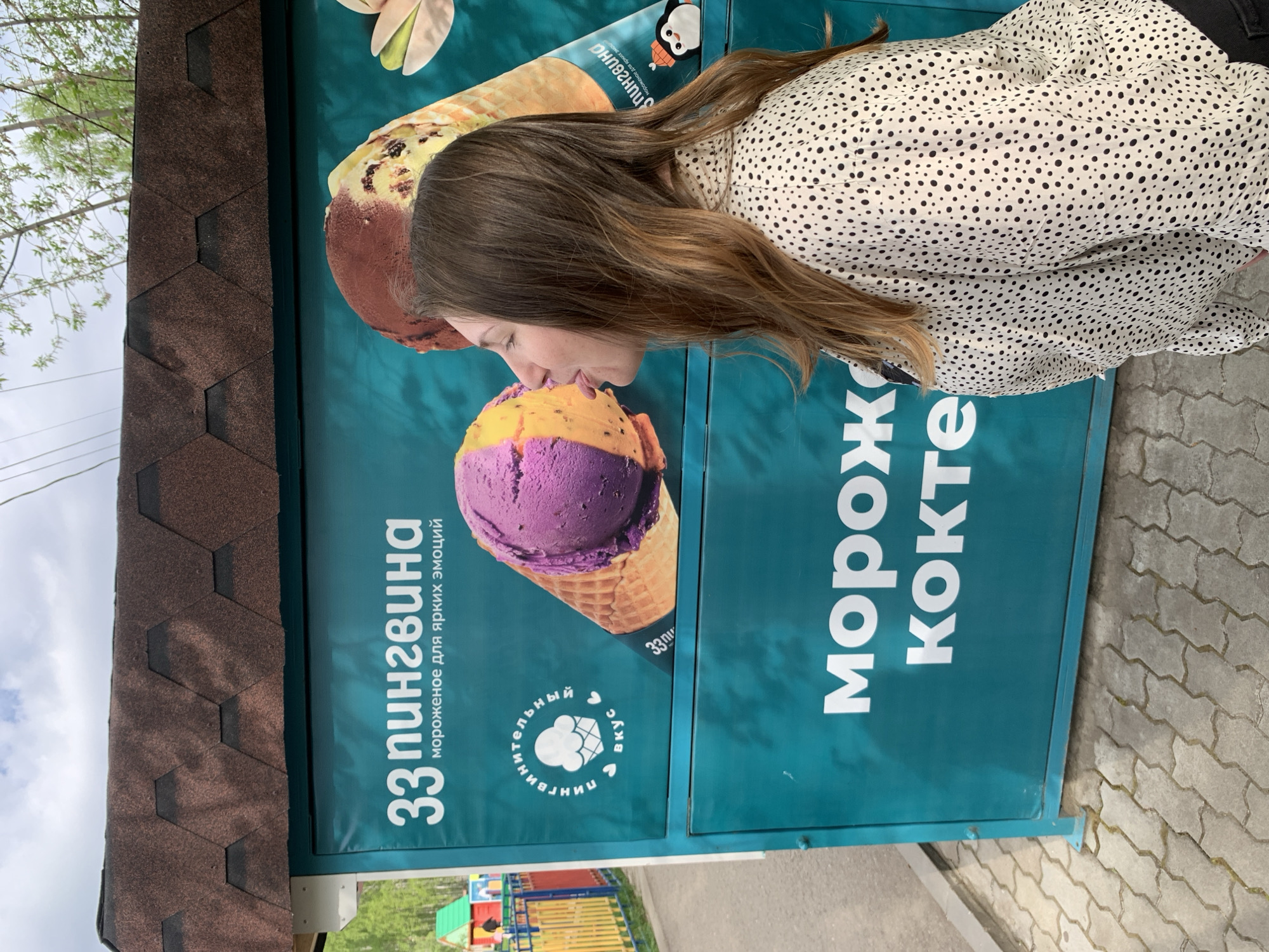 The image shows a woman appearing to lick a large advertisement for "33 пингвина" ice cream, with the words "морож кокте" (ice cream cocktail) written on the side. The ad features images of ice cream cones, one with chocolate and vanilla ice cream, and another with purple and orange ice cream. The location is in Chaikovsky, Russia. The woman is wearing a white shirt with black polka dots. A building with a brown shingle roof is visible behind the sign, and a children's playground is in the background. The ground is paved with interlocking concrete blocks.