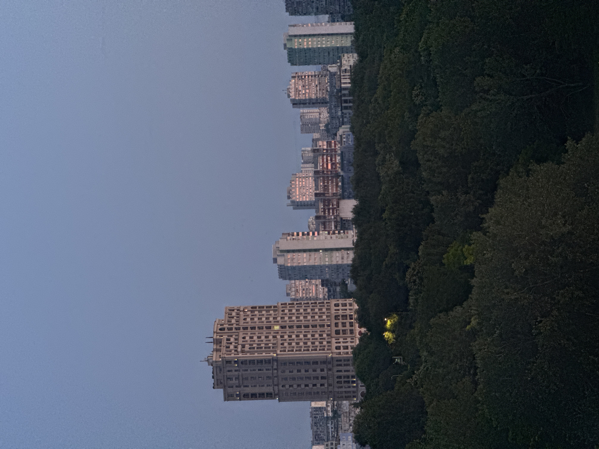 This image depicts a cityscape at dusk, with a foreground of dark, lush trees partially obscuring a backdrop of tall buildings. The sky is a clear, pale blue, indicating the transition from day to night. In the mid-ground, a prominent, ornate apartment building with many windows stands out, its light beige facade detailed with architectural features. To its right, several other apartment buildings of varying heights and architectural styles are visible, some with windows illuminated by warm, orange light. Further in the distance, more buildings form a dense urban silhouette. The trees in the foreground are dark and dense, with a few streetlights casting a yellow glow through the foliage. The overall scene captures a twilight view of an urban environment, likely a park or green space bordering a residential area.
The image was taken in Burgas, Bulgaria, from a high vantage point, likely an apartment building. The architecture of the buildings, particularly the prominent beige apartment block, is characteristic of some residential areas in Burgas. The presence of extensive greenery in the foreground suggests a location near a park or a forested area within or bordering the city.