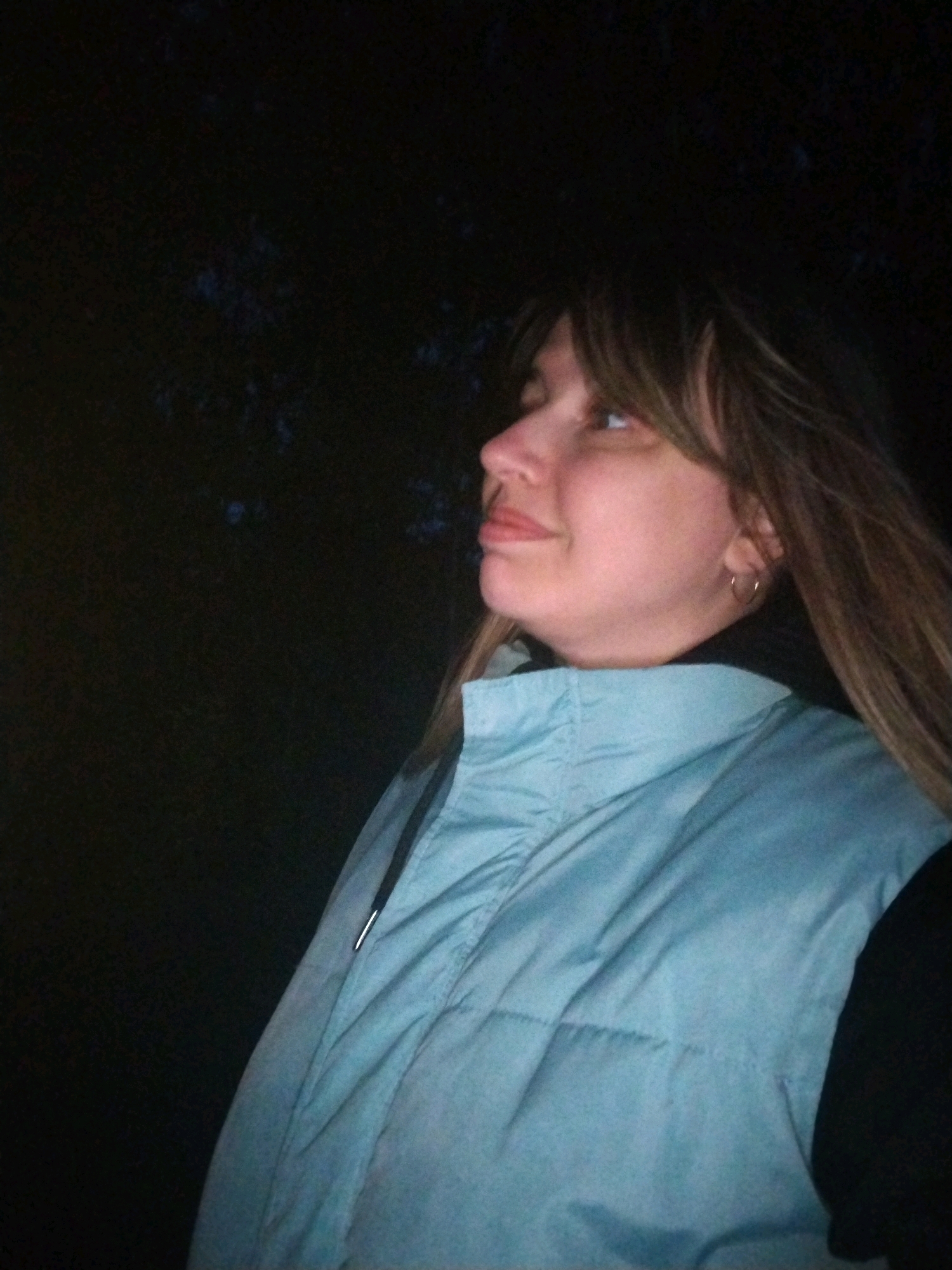 A medium shot captures a young woman from the chest up, looking upwards and to the left. She has long, light brown hair with bangs and is wearing a light blue puffer vest over a black garment. The background is dark and appears to be outdoors, with blurred trees or foliage visible. The lighting suggests it is nighttime or a very dimly lit environment. Her facial features are visible, with a slight smile and an earring in her visible ear. No specific location within Yaroslavl, Russia, can be identified from the image.