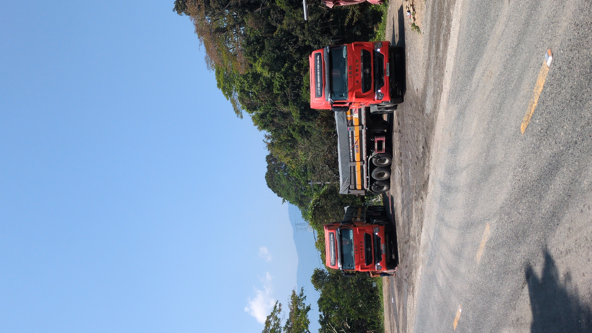 The image captures two red trucks parked on an asphalt road, partially obscured by trees. The scene is set in Lâm Sơn, Vietnam, as indicated by the "CTY CỔ PHẦN KHOÁNG SẢN & VLXD LÀM ĐỒNG" (joint stock company for minerals and construction materials), a business located in the area. The sky is a clear blue. A yellow line is painted on the road. The trucks are parked on the side of the road. The image is oriented sideways. A mountain is visible in the background.