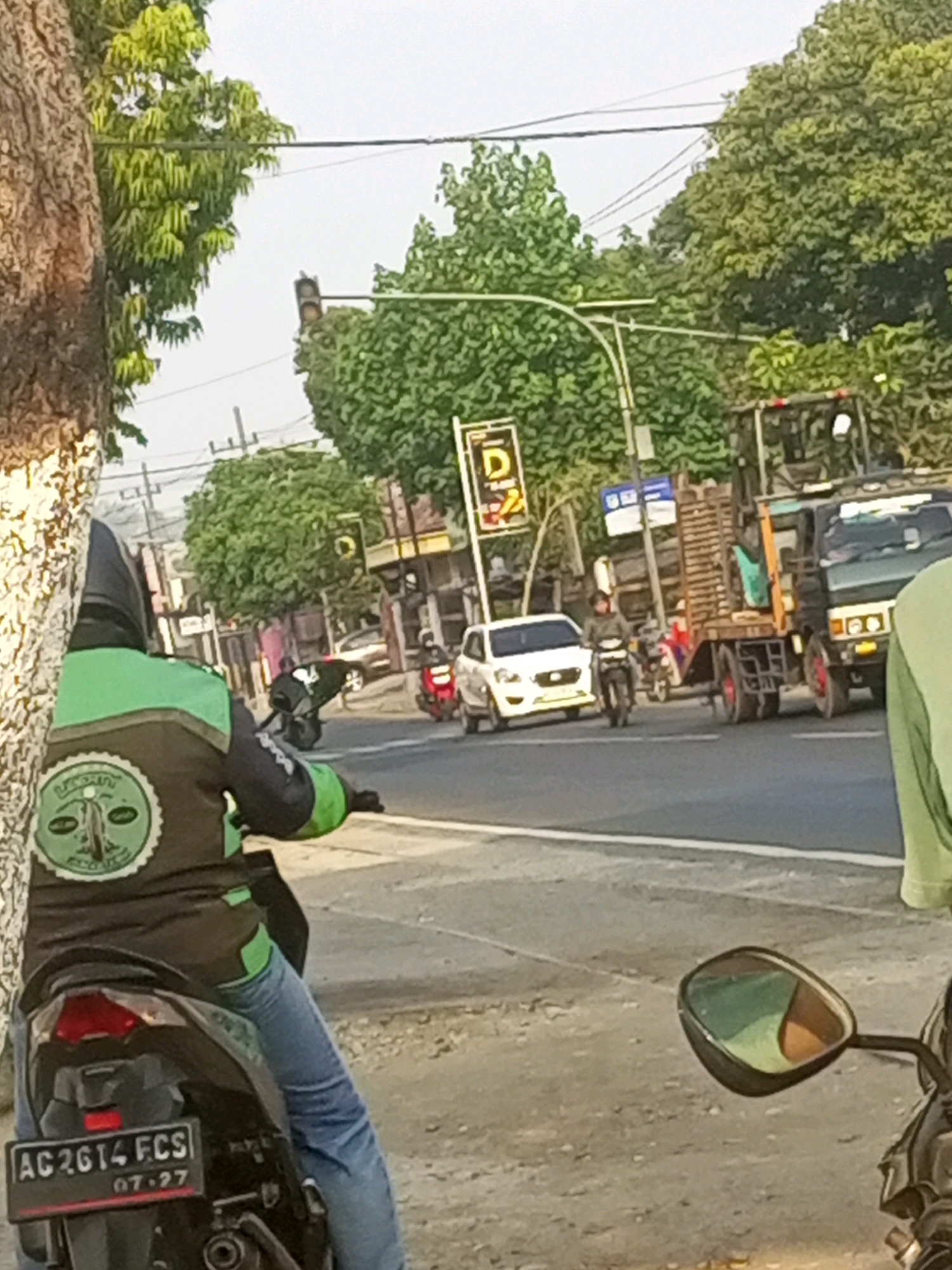 The image captures a street scene in Gurah, Indonesia, with a focus on a person riding a motorcycle in the foreground. The rider is wearing a black jacket with green accents and a circular emblem on the back, and blue jeans. The motorcycle's license plate reads "AC 2614 FCS" with "07-27" below it.
In the background, a white car is visible on the road, as is a truck carrying wooden planks. Further back, traffic lights and trees line the street, with buildings suggesting an urban or semi-urban environment. The overall lighting suggests it is daytime, possibly late afternoon given the soft light. The scene appears to be an ordinary moment of traffic and daily life in the depicted location.