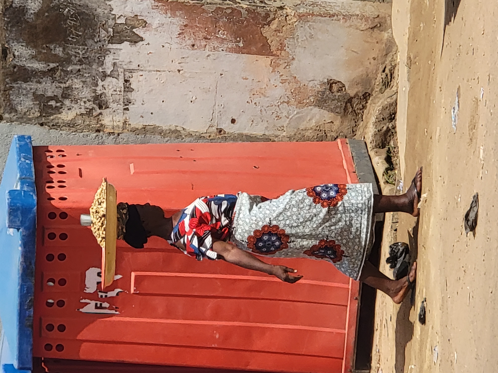 The image captures an outdoor scene in Maiburiji, Nigeria, bathed in bright, clear sunlight. The central subject is a person, likely a woman, standing and expertly balancing a flat, rectangular wooden tray filled with groundnuts (peanuts) on her head. She is dressed in traditional African attire, consisting of a vibrant, multi-patterned blouse featuring red, blue, and white geometric designs, paired with a light-colored skirt adorned with large, symmetrical blue and orange floral motifs.
Her feet are bare on the sandy-brown dirt ground; one black flip-flop lies beside her foot. Behind the woman is a substantial, corrugated red structure, possibly a kiosk, container, or part of a building, with a series of small, recessed circular shapes and horizontal lines. A portion of a blue structure is visible to the left, appearing to be connected to the red one. The background features a weathered wall with peeling white paint, revealing underlying darker tones and textures. The strong sunlight indicates a daytime setting. No clear, legible text is visible on any of the surfaces. The scene depicts a common sight of street vending or transportation of goods.