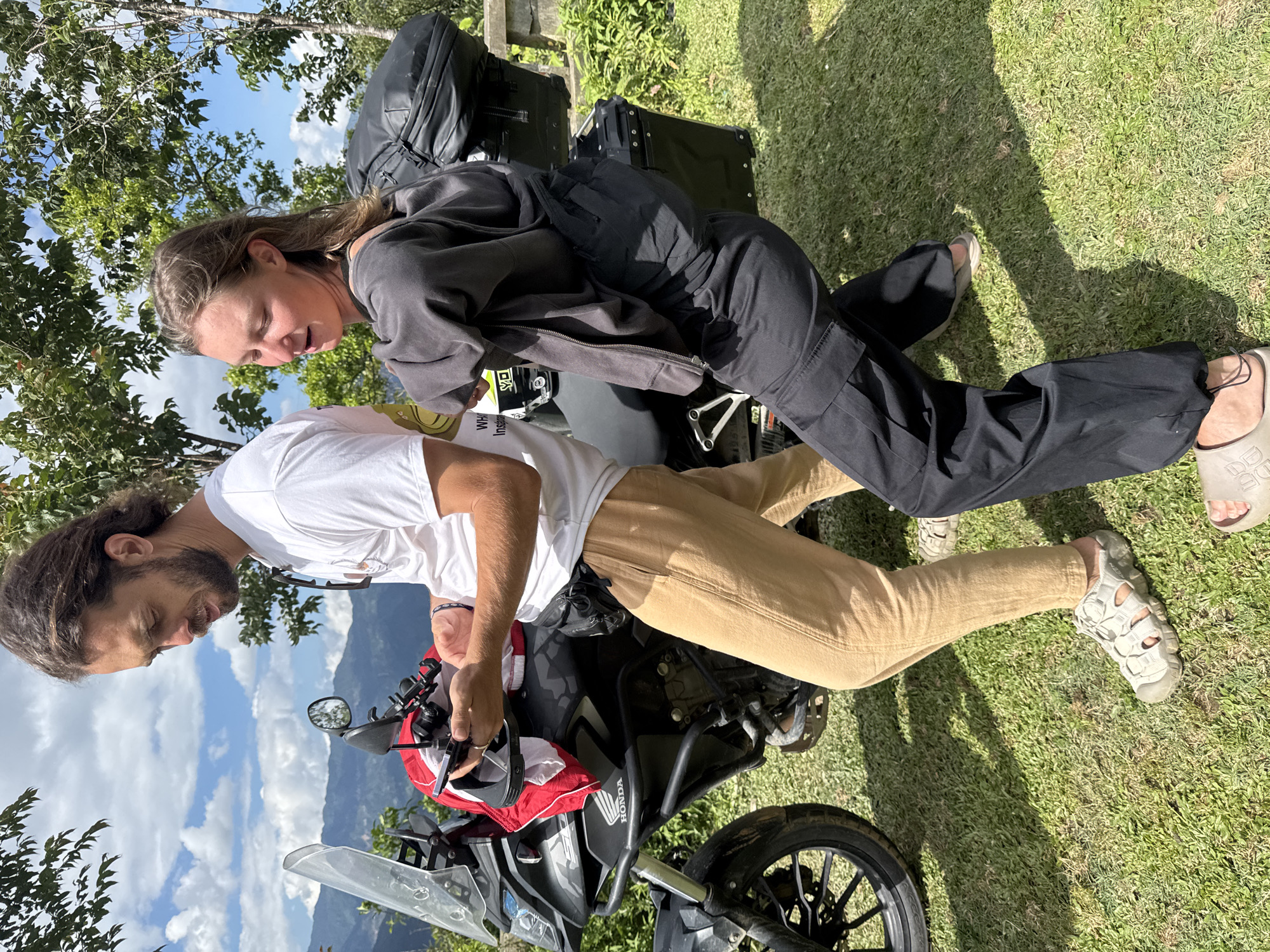 An outdoor scene in Tu Le, Vietnam, features a man and a woman standing on a grassy patch beside a parked black Honda adventure motorcycle, set against a backdrop of distant green mountains and a partly cloudy blue sky.
The man, positioned on the left, is wearing a white short-sleeved t-shirt with a partial "WH" graphic, light beige trousers, and grey closed-toe sandals. A black fanny pack is visible at his waist, and sunglasses hang from his shirt collar. He has dark, shoulder-length hair and a beard, and is intently looking down at and manipulating a smartphone secured in a mount on the motorcycle's handlebars. His hands are focused on the device.
The woman, positioned on the right, wears a dark grey or black long-sleeved hooded jacket, black loose-fitting cargo trousers with elasticized cuffs, and light-colored slip-on sandals. Her light brown hair is pushed back. She is oriented towards the motorcycle, with her head tilted back and to her right, and her eyes partially closed, seemingly in motion or adjusting her posture. A large black soft-sided bag is strapped to the rear of the motorcycle, directly behind her.
The black Honda motorcycle, possibly a CB500X, is equipped with a clear windshield, black crash bars, and black hard-shell panniers on the rear, in addition to the soft-sided bag. A red and white item is draped over the handlebars near the man's hands. The ground beneath them is covered in short, vibrant green grass, casting distinct shadows indicating bright sunlight from above. Green trees with dense foliage are visible behind the individuals, further defining the natural, outdoor setting.