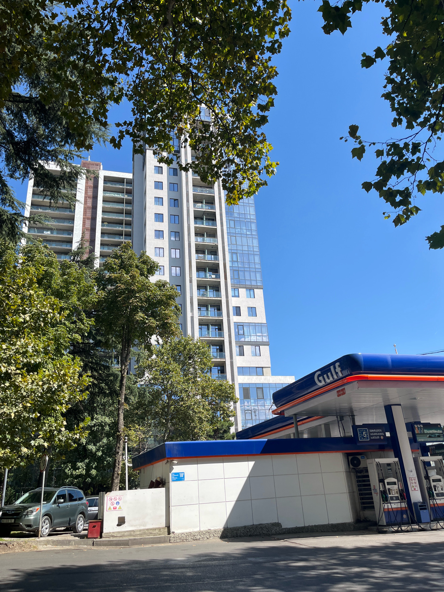 The image depicts a clear blue sky above a modern apartment building and a Gulf gas station in Tbilisi, Georgia. Trees with green leaves are visible in the foreground and frame the scene. The apartment building is tall and sleek, with a glass facade and balconies. The gas station has a blue and orange canopy with the "Gulf" logo. Several cars are parked nearby, including a dark green SUV on the left. The environment appears to be an urban setting with well-maintained infrastructure.