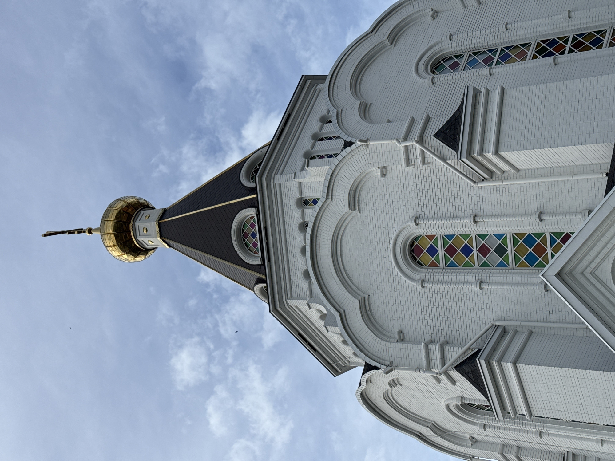 The image depicts the exterior of a building, possibly a church or cathedral, viewed from a low angle looking upwards towards the sky. The building is predominantly white with brickwork visible on the surface. It features arched windows filled with stained glass composed of small, colorful diamond-shaped panes. The architecture includes decorative moldings and cornices, suggesting a classical or Byzantine influence. Above the main structure, there is a dark-colored roof with a pointed dome topped by a golden onion-shaped cupola and a cross. The sky is light blue with scattered clouds. The location is Unknown, Unknown.