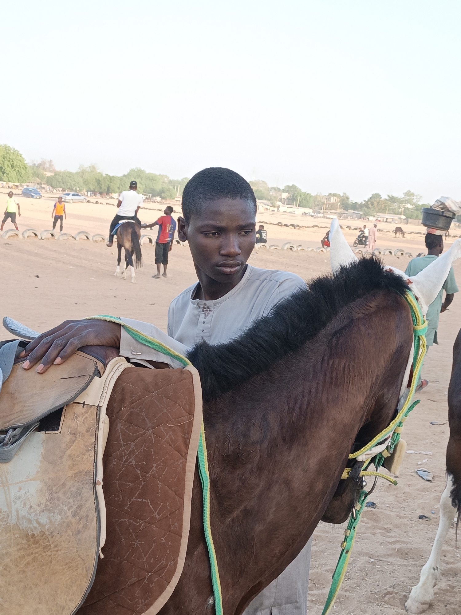In Giginyu, Nigeria, a young man in a light-colored, buttoned shirt stands next to a saddled brown horse with white ears. The horse has a black mane and green and yellow rope reins. The man’s left hand rests on the saddle. In the background, several people are gathered; one is riding another horse, and another is carrying a container on his head. A line of tires marks a boundary. The ground is sandy.