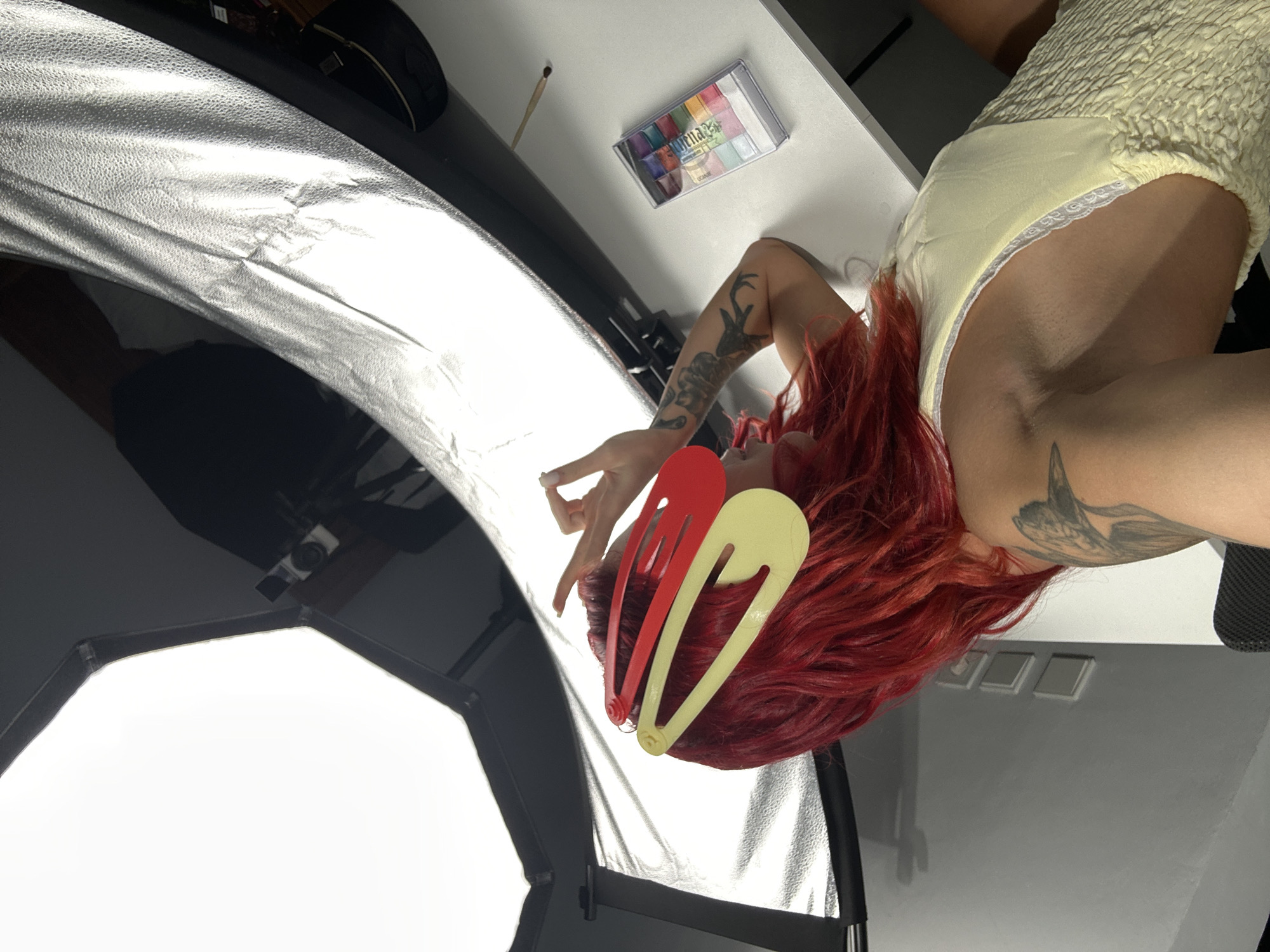 In North Kuta, Indonesia, a person with bright red hair, styled with a red and yellow hair clip, is seen from behind. They are wearing a yellow smocked dress with lace trim. The person is leaning forward, with their left arm extended and their hand resting on their head. Their right arm is visible, showing tattoos. In the background, a large photographic softbox is positioned overhead, casting bright light. To the right, a white table holds a palette of colorful eyeshadows and a slender stick, possibly a makeup brush. The environment appears to be a studio or well-lit indoor space.