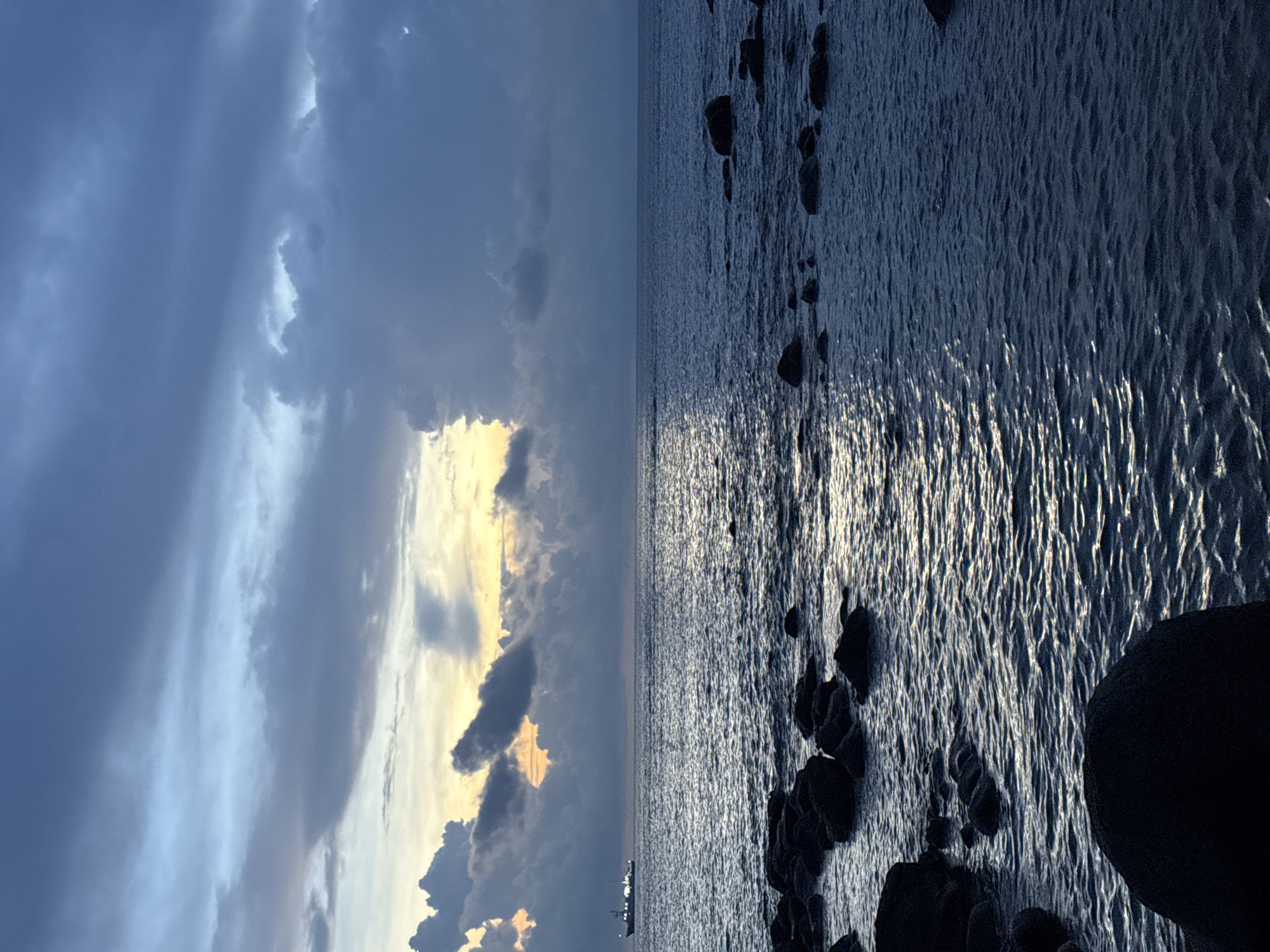 An image captures a serene seascape at dusk or dawn, featuring a vast expanse of dark blue ocean meeting a dramatic sky. The sky is a canvas of swirling clouds, with patches of soft orange and yellow light peeking through from the horizon, suggesting the sun is either rising or setting. In the distance, a lone cargo ship is visible on the calm water, its silhouette dark against the illuminated sky. The foreground is composed of dark, jagged rocks of varying sizes partially submerged in the water, creating a rugged shoreline. The water itself is rippled, reflecting the muted colors of the sky, with streaks of light shimmering on its surface. The scene is one of natural beauty and tranquility, likely situated on the coast of Lý Sơn Special Administrative Region, Vietnam, given the characteristic rocky shoreline and the type of vessel common in the region. There are no people or animals visible in the image.