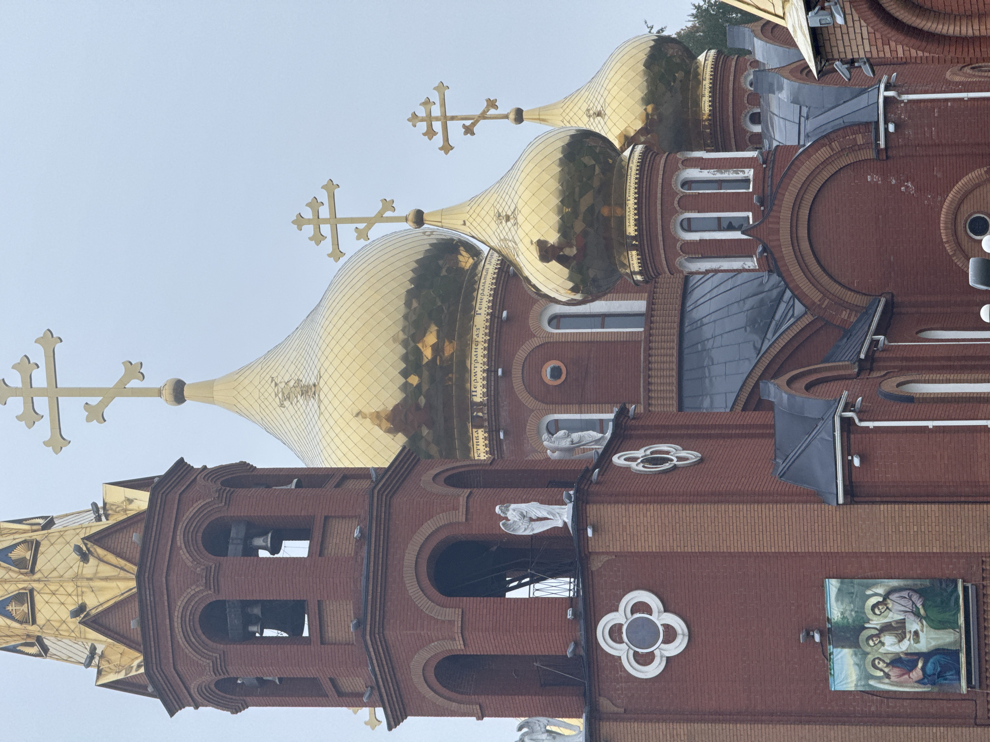 This image captures a vibrant, multi-domed Eastern Orthodox church, presented in a portrait orientation that has been rotated 90 degrees clockwise from its natural upright position. The church features a striking facade constructed primarily of red brick, accented with lighter beige bricks forming intricate archways and decorative patterns.
Dominating the upper section are several gleaming golden onion domes, each crowned with an ornate Orthodox cross. The highly reflective surfaces of these domes show visible Cyrillic text, including phrases such as "ГЕНЕРАЛЬНЫЙ ДИРЕКТОР" (General Director) and the city name "ПЕРМЬ" (Perm). This text appears to be reflections from surrounding elements rather than direct inscriptions on the domes.
The church complex includes a prominent bell tower with arched openings, through which large bells are partially visible. Adorning the brickwork are several white sculptures, possibly depicting angels or saints, and decorative white clover-like motifs. A large rectangular religious icon or painting, depicting figures with halos, is affixed to the exterior wall.
The scene is set under an overcast or hazy sky, indicating daytime with diffuse lighting, as no harsh shadows are apparent. The architectural style, particularly the onion domes and Orthodox crosses, along with the visible Cyrillic text, firmly places the location in a Russian-speaking region, specifically Chaikovsky, Russia. No specific activity or event is taking place; the image is a static portrayal of the church's exterior.