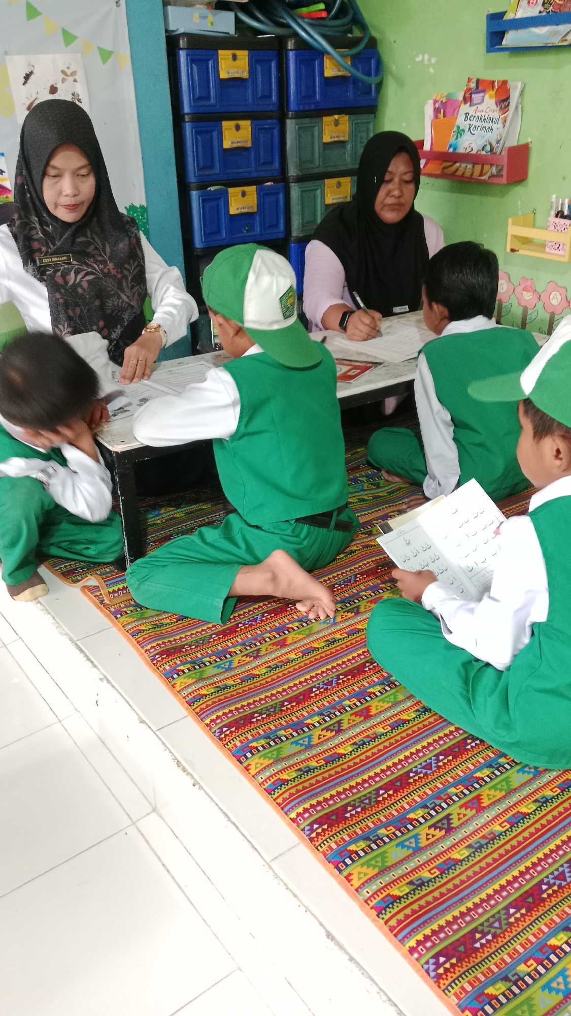 In a classroom setting, likely in Karangharjo, Indonesia, a group of young children, dressed in green uniforms and caps, are engaged in learning activities. Two adult women, presumably teachers, are also present. One teacher, wearing a black hijab and white blouse with a patterned scarf, leans over a table, her attention focused on a student. The other teacher, also in a black hijab, sits at the table, writing on a paper. The children are seated on a colorful mat on the floor around the table. Some are actively participating, looking at books or writing, while one child appears to be resting his head on the table. The room is decorated with educational materials, including books on shelves and colorful wall art. Storage units with multiple drawers are visible in the background, suggesting an organized classroom environment. The overall scene depicts a typical day of learning and interaction in a kindergarten or early primary school.