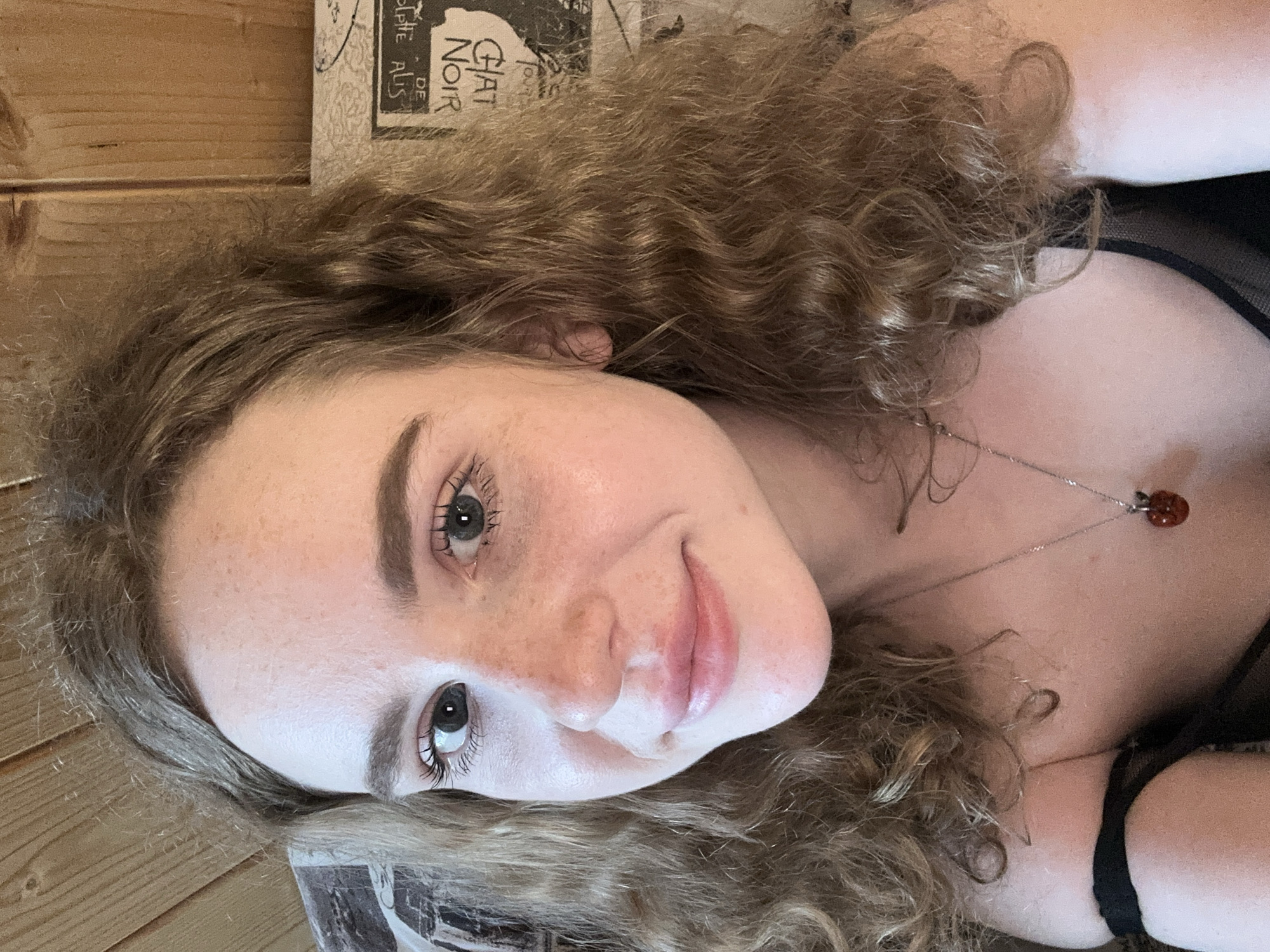 A close-up portrait shows a young woman with long, wavy, light brown hair. She lies down, looking directly at the camera with a gentle, soft smile and slightly parted lips. Her skin features visible freckles across her nose and cheeks, complementing her light-colored eyes. She wears a delicate silver-colored necklace with a small, reddish-brown pendant, possibly amber or a ladybug charm. A dark, possibly sheer, top is visible on her upper body.
The background on the left side of the frame features light-colored wooden planks, suggesting a wall or furniture. On the right, partially visible, is printed material, likely a poster or book cover. Clearly discernible text on this item includes "CHAT NOIR" and "DE SOLPIE A.LIS," referencing the historical "Le Chat Noir" cabaret. The lighting is soft and natural, contributing to a serene atmosphere. The image captures a moment of quiet repose, likely taken indoors in Vasilyevo, Russia.