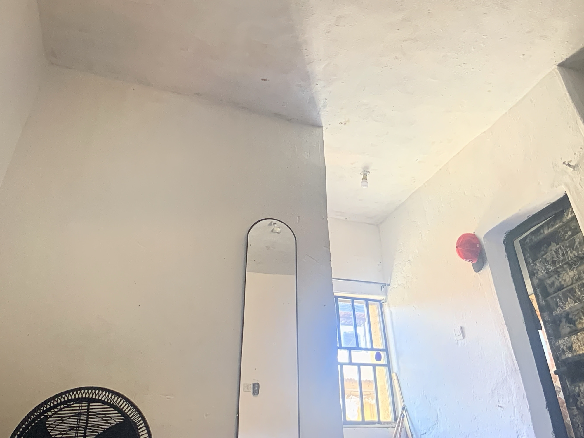 This is an interior, upward-angled shot of a room or hallway, characterized by off-white, textured walls and ceiling. The scene is illuminated by natural light entering through a window and supplemented by an incandescent light bulb fixture on the ceiling.
In the center-left, a tall, arched, floor-length mirror reflects the opposite wall and ceiling. To its left, the top portion of a black oscillating fan is visible at the bottom edge of the frame. The walls show signs of wear and uneven texture, suggesting plaster or a similar material. A distinct horizontal line is visible across the ceiling and upper wall sections, where the texture or shade changes, possibly indicating a beam, a new paint application, or a different construction material.
On the right side of the image, a large window with metal bars or a grid pattern allows daylight to stream in, revealing an exterior scene that is partially obscured but appears to be another building or structure. Leaning against the wall near the window is a thin, light-colored pole or stick. High on the wall to the right of the mirror, a red object resembling a hard hat or construction helmet is hanging. Further to the right, a dark, possibly patterned door is partially visible, appearing to be set within a thick archway. A white light switch or outlet is visible on the wall between the mirror and the window.
No people are present in the image, and no specific activity is taking place. The room appears to be a functional space, possibly an older dwelling or one that is undergoing some work, given the textures and the visible objects. No text is visible in the image.