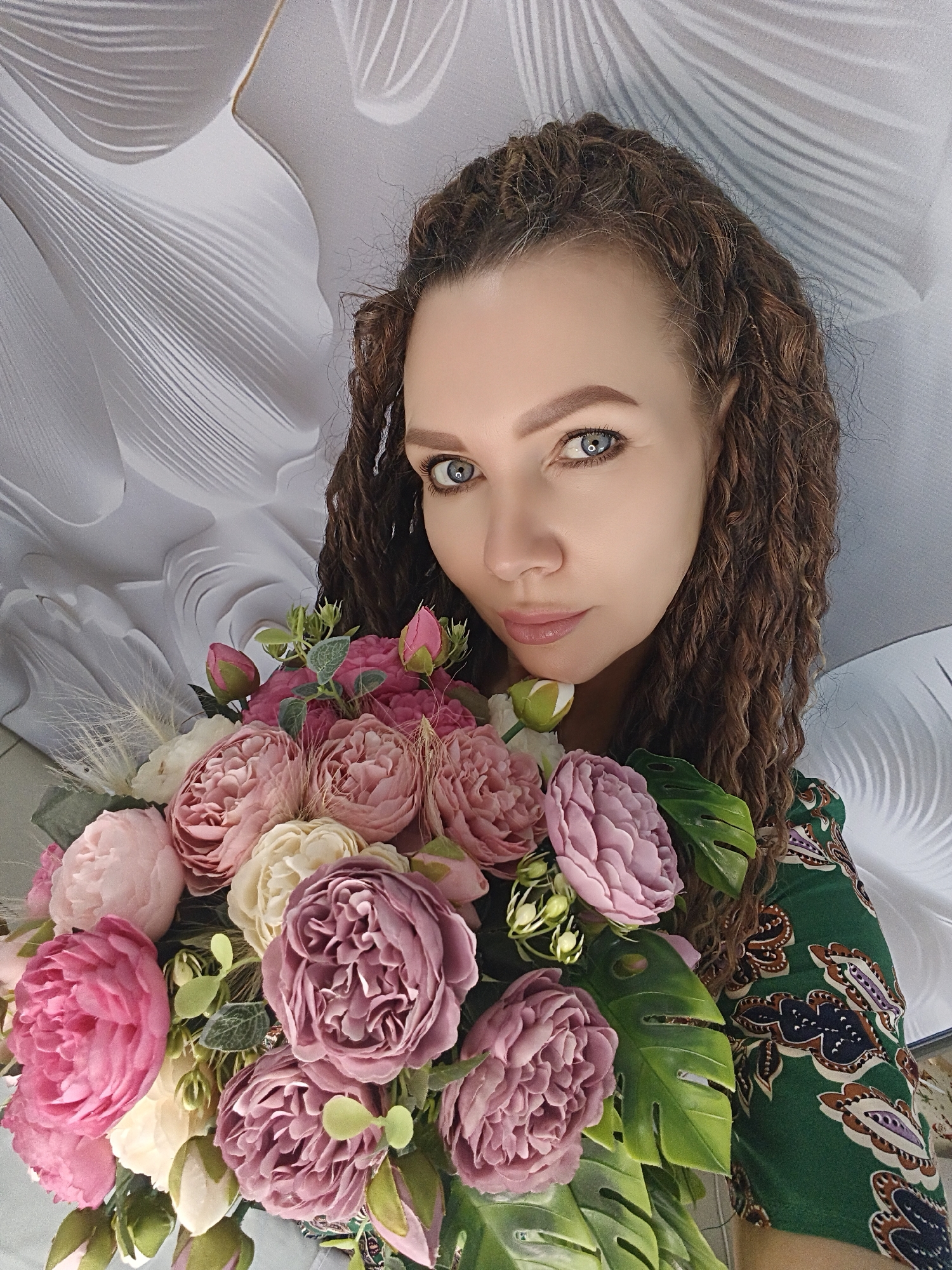 A woman is holding a bouquet of flowers. The woman has brown curly hair and blue eyes. She is wearing a green paisley print dress. The bouquet contains pink and white peonies, along with greenery including monstera leaves. The background features a textured white and gray pattern, possibly a wall or ceiling. The location is unidentified.