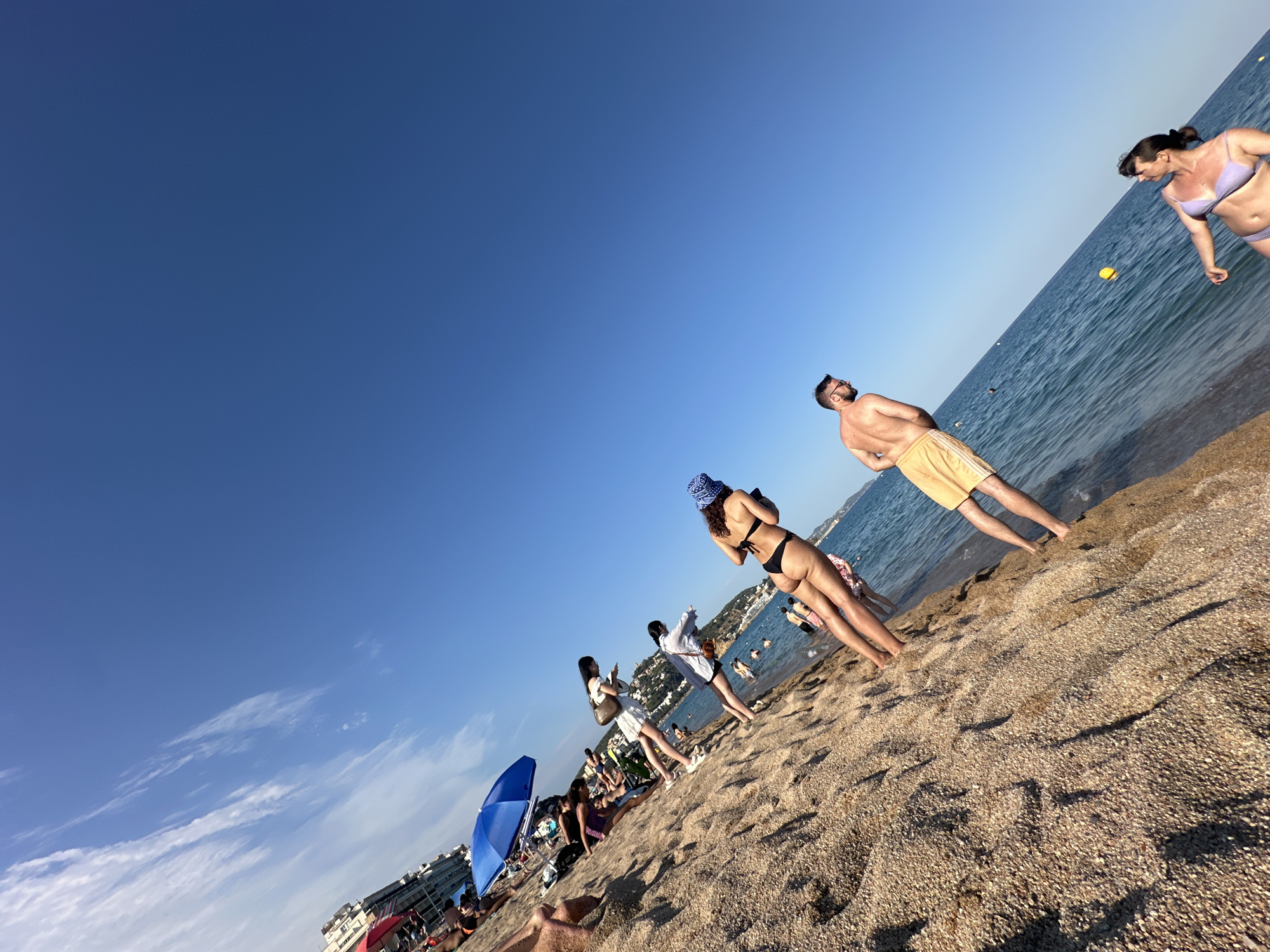 A group of people are on a sandy beach in Blanes, Spain, with the Mediterranean Sea visible. The sky is clear and blue with some wispy clouds. On the left side of the frame, several people are seated or standing under a blue umbrella, with buildings in the background. Further into the scene, closer to the water, three women are walking on the sand. One woman in the foreground is wearing a black bikini and a blue hat, and is facing away from the camera. To her left, two other women are walking in the same direction. In the middle ground, a man in yellow shorts is standing in the shallow water, facing a woman in a purple bikini who is in the lower right corner of the frame, also in the water. Several other people are visible in the water in the distance.